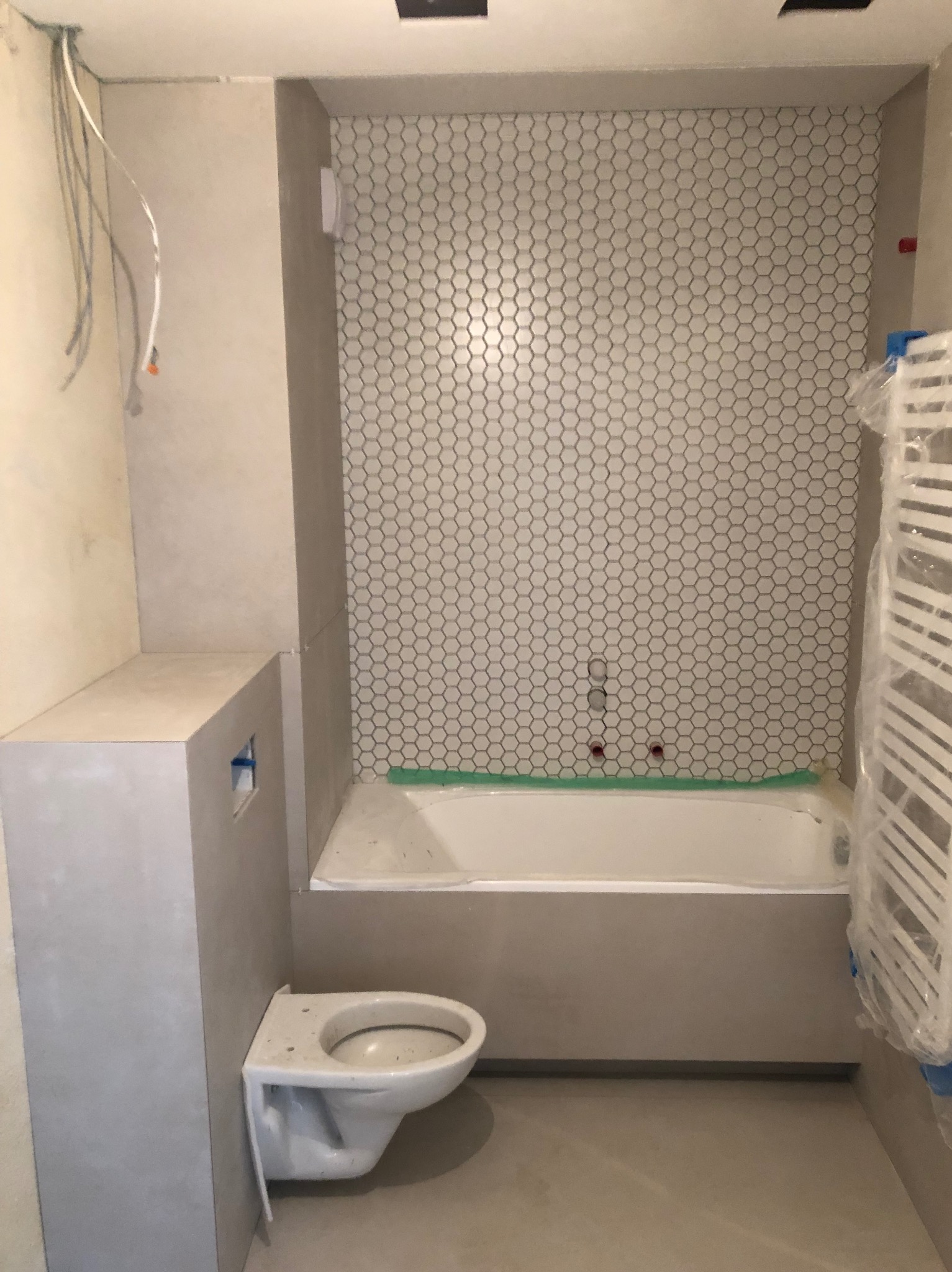 Niedokończona łazienka z wanną, płytkami heksagonalnymi na ścianie, podwieszaną toaletą w zabudowie i grzejnikiem drabinkowym w folii ochronnej; widoczne wystające przewody elektryczne.