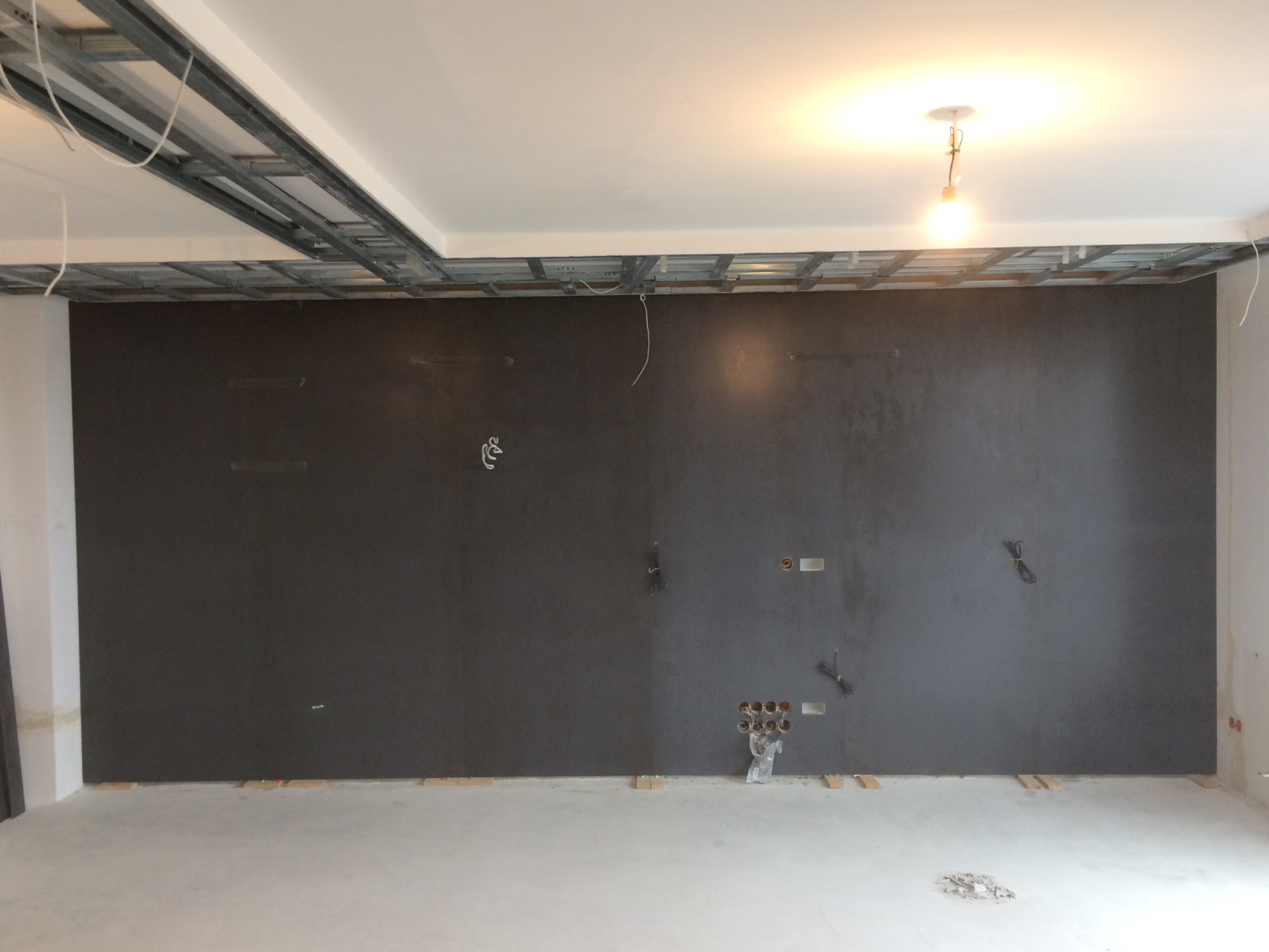 Ściana pomalowana na ciemny kolor w trakcie wykończenia wnętrza, widoczne wystające kable, puszki elektryczne, profile sufitowe i drewniane podkładki pod ścianą.