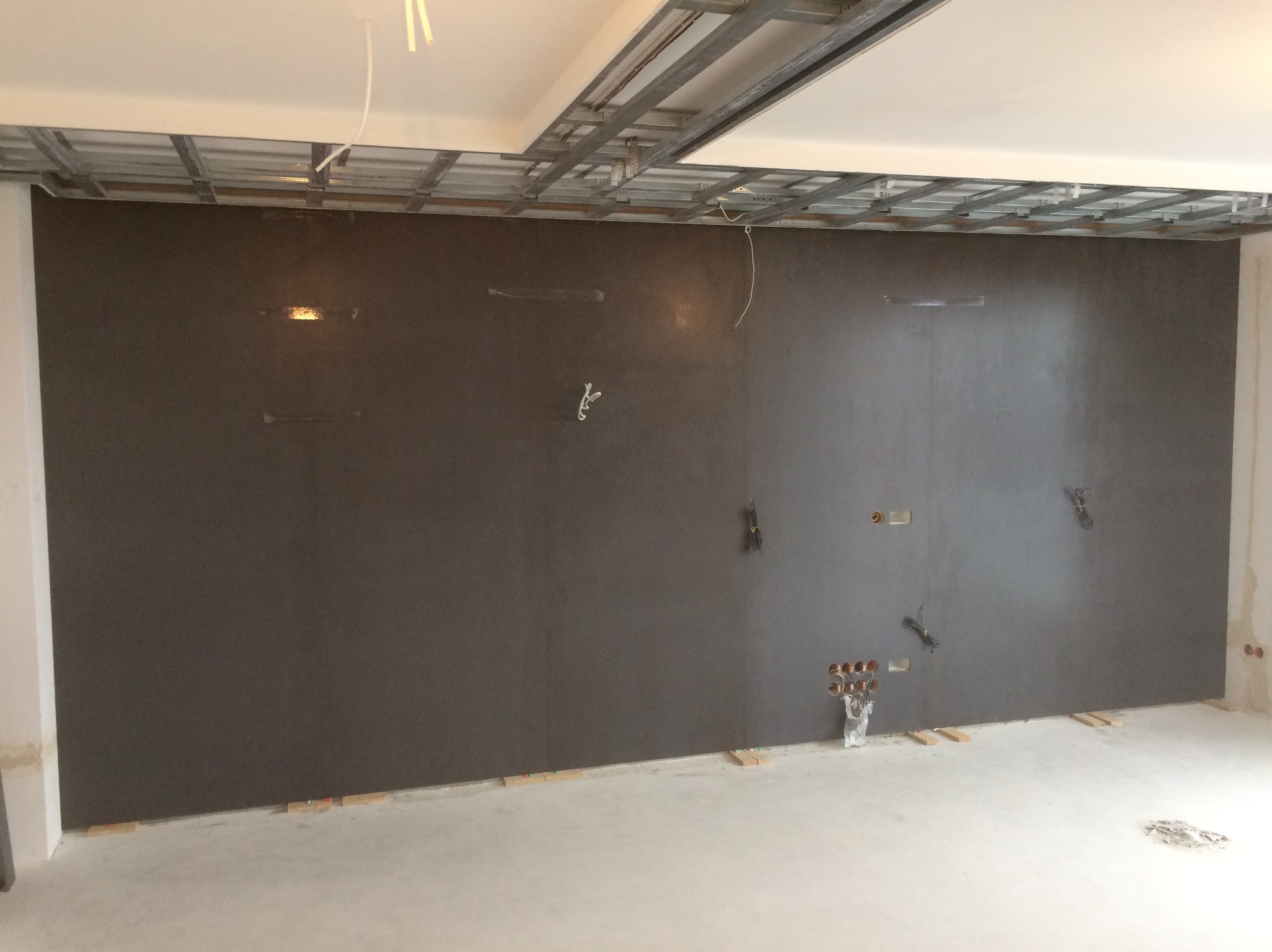 Szara ściana z instalacją elektryczną i wodną w surowym stanie, widoczne drewniane podkładki pod ścianą i metalowy stelaż pod sufitem podwieszanym.