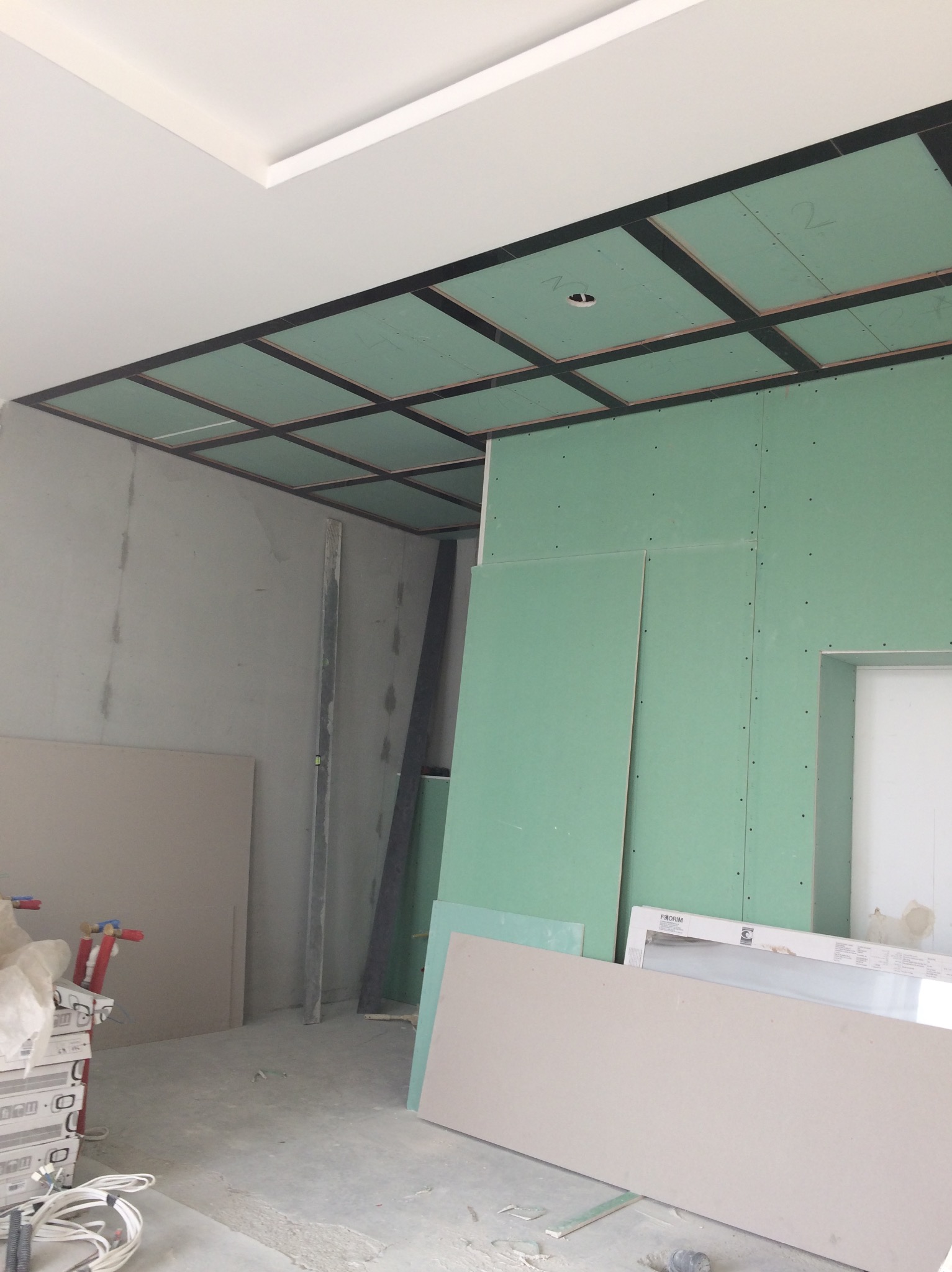 Wnętrze w trakcie prac wykończeniowych: zielone płyty gipsowo-kartonowe na ścianach i suficie z kratownicą, widoczne materiały budowlane i narzędzia w nieuporządkowanym otoczeniu.
