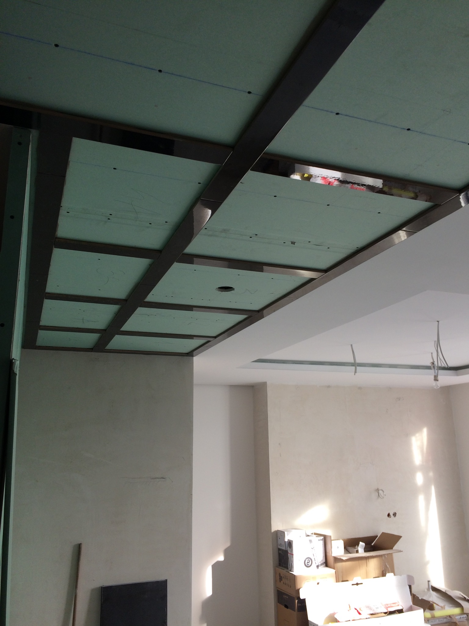 Sufit podwieszany z zielonej płyty gipsowo-kartonowej, widoczne stalowe profile, w tle surowe ściany i kartony z materiałami budowlanymi.