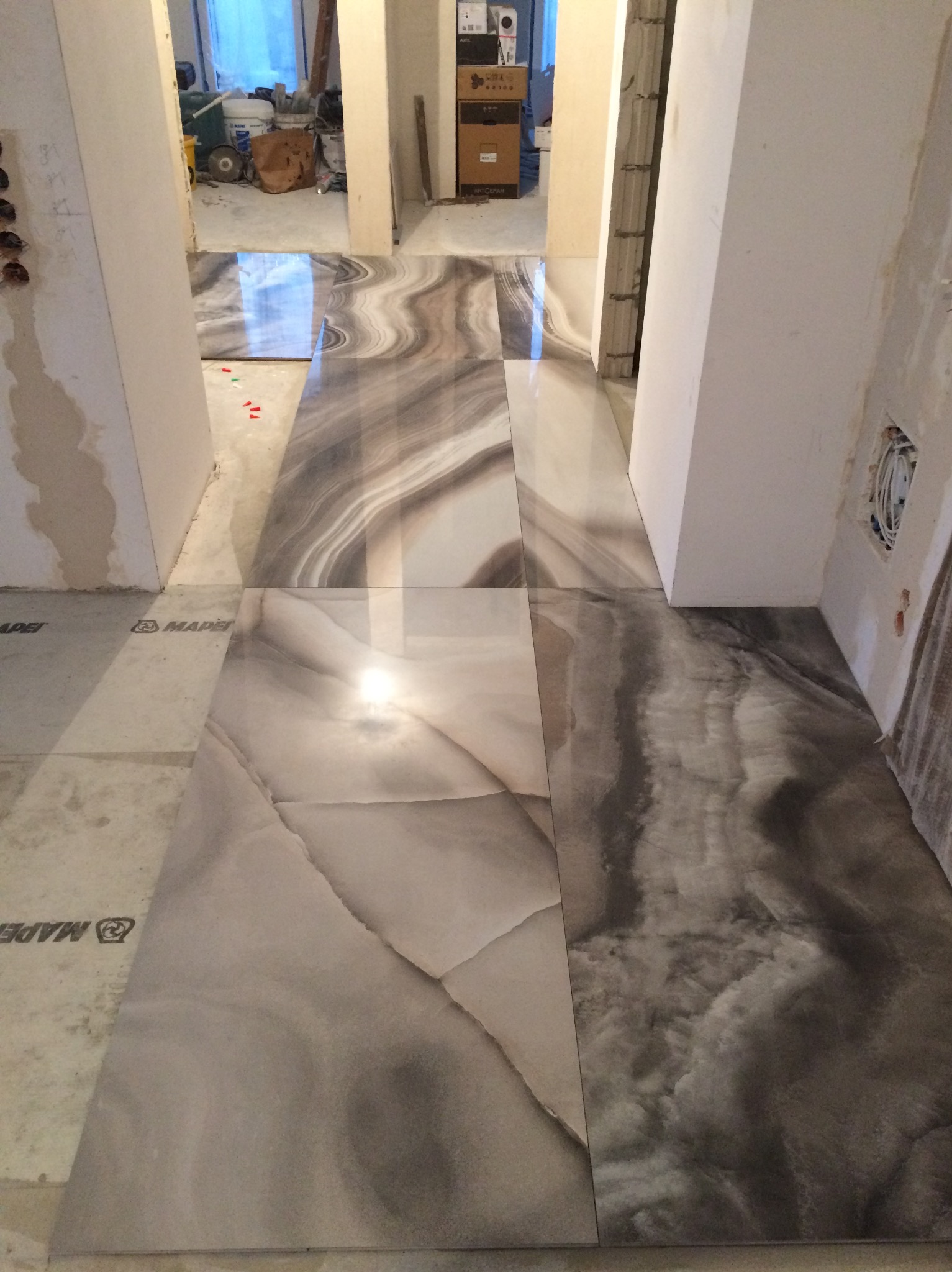 Układanie błyszczących płytek podłogowych imitujących marmur w odcieniach szarości i beżu, widoczne niedokończone ściany i materiały budowlane w tle.