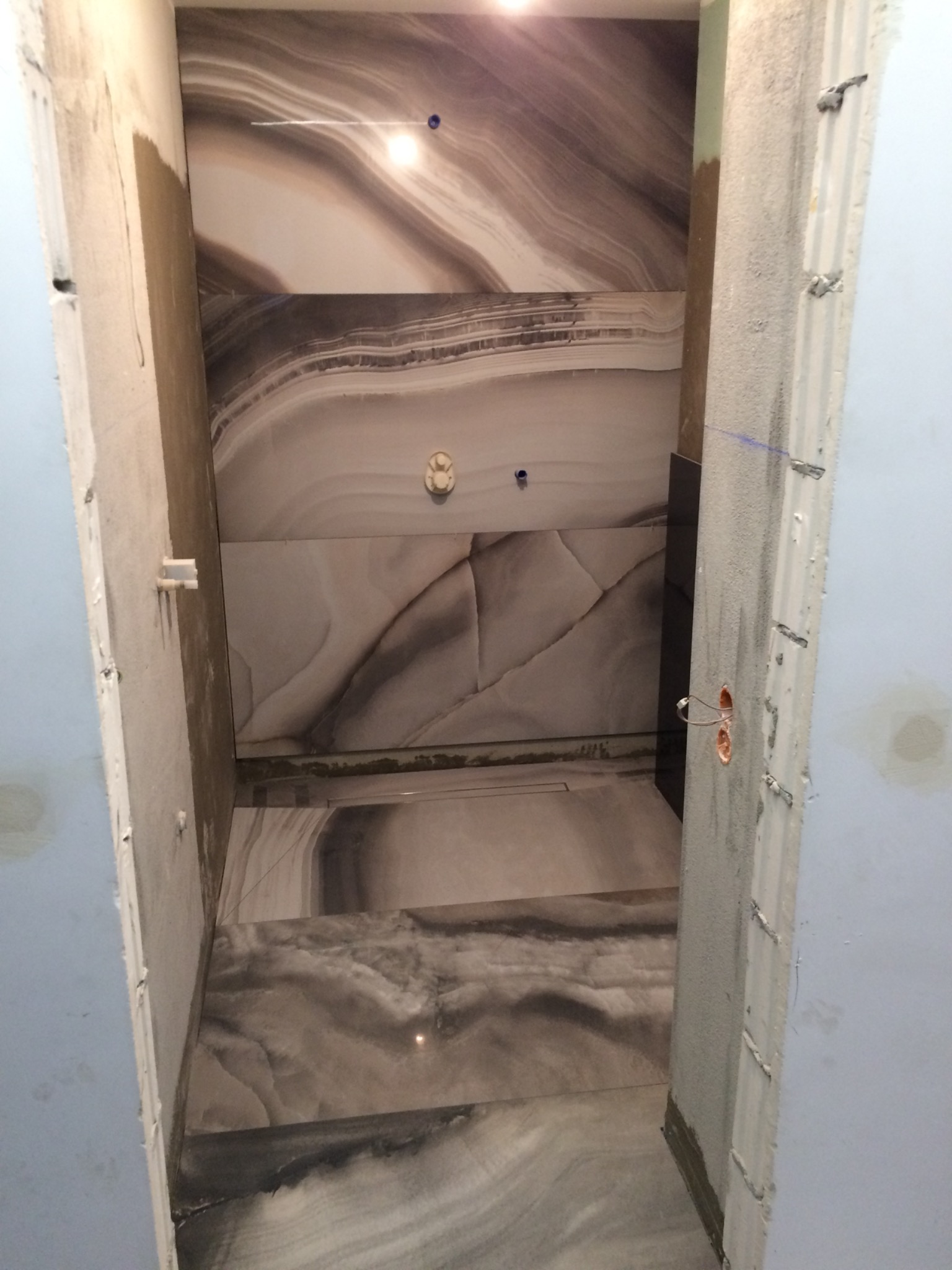 Wnętrze łazienki w trakcie prac wykończeniowych: położone płytki imitujące marmur na ścianach i podłodze, surowy tynk na framudze drzwi.