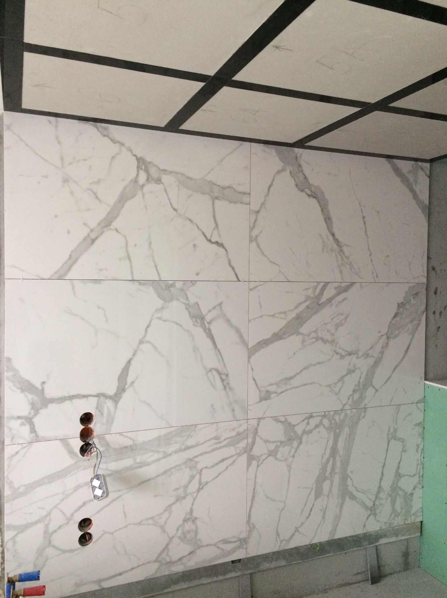 Ściana wyłożona wielkoformatowymi płytkami imitującymi marmur, widoczne otwory pod instalację elektryczną i fragment sufitu z zamontowaną konstrukcją z profili.