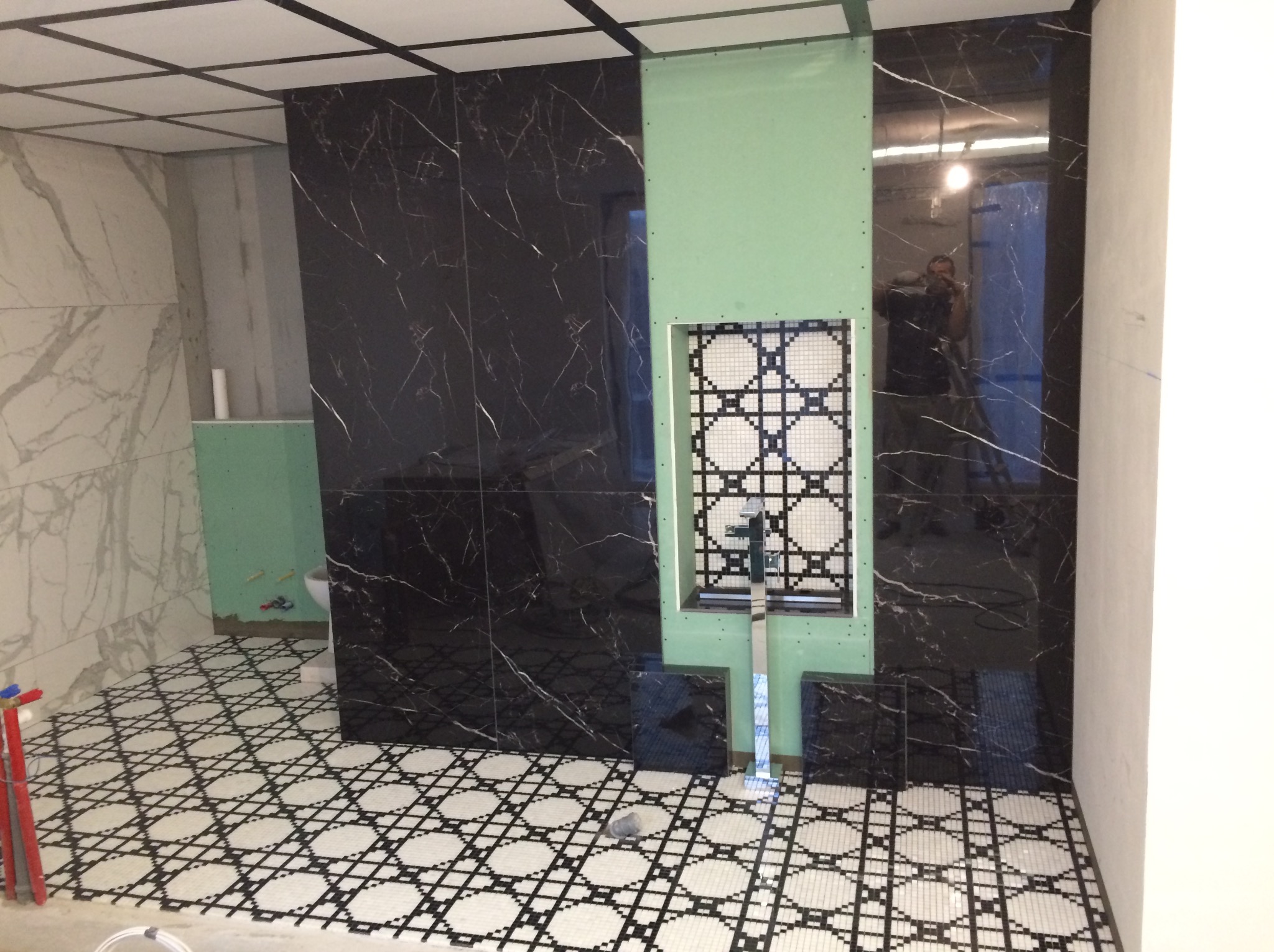 Łazienka w trakcie wykańczania: czarne płytki imitujące marmur na ścianach, mozaika w niszy obudowanej płytą kartonowo-gipsową, wzorzysta podłoga, widoczne narzędzia i odbicie fotografa...