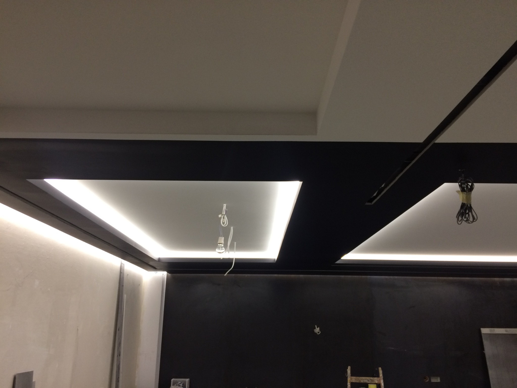 Sufit podwieszany z oświetleniem LED, czarne elementy dekoracyjne, widoczne kable i oprawki lamp podczas montażu, surowa ściana w tle.