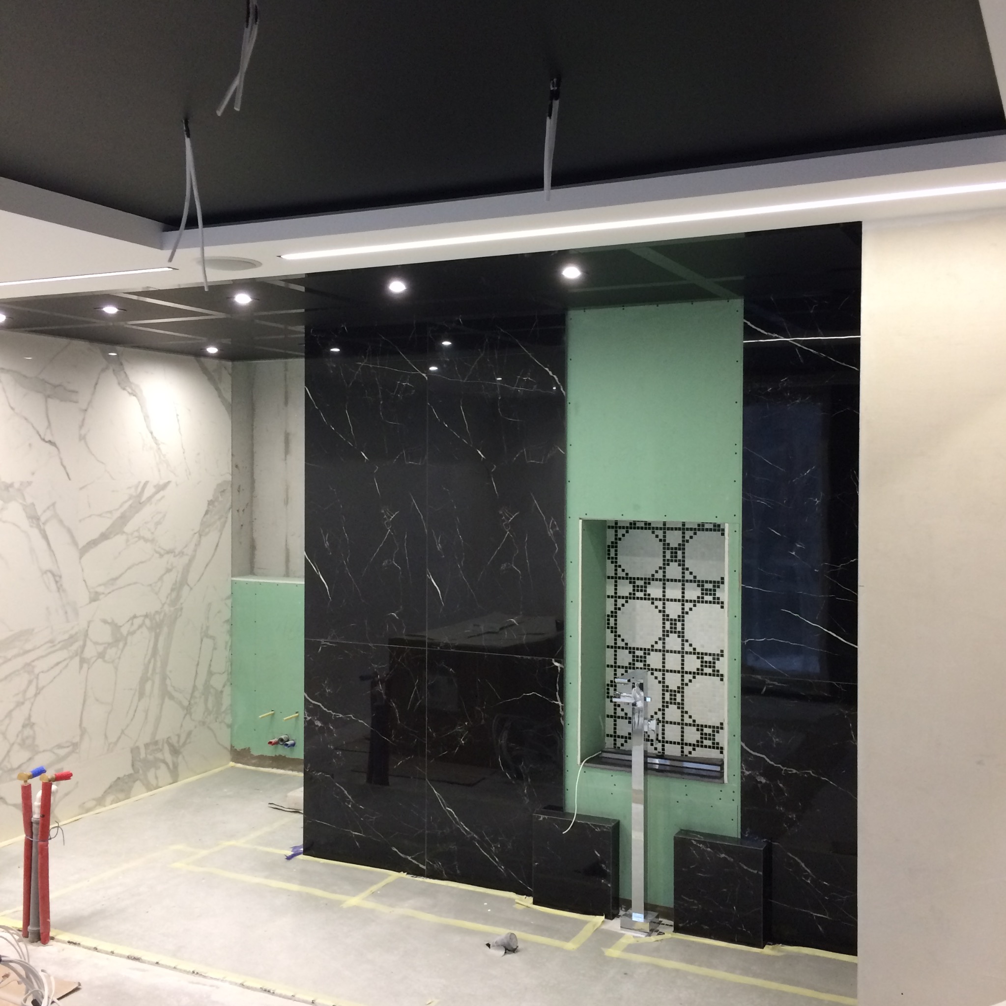 Wnętrze w trakcie prac wykończeniowych: czarne płytki imitujące marmur na ścianie, fragment ściany z płyty karton-gips z wyciętym otworem na dekoracyjną mozaikę, widoczne rury instalacji wodnej...