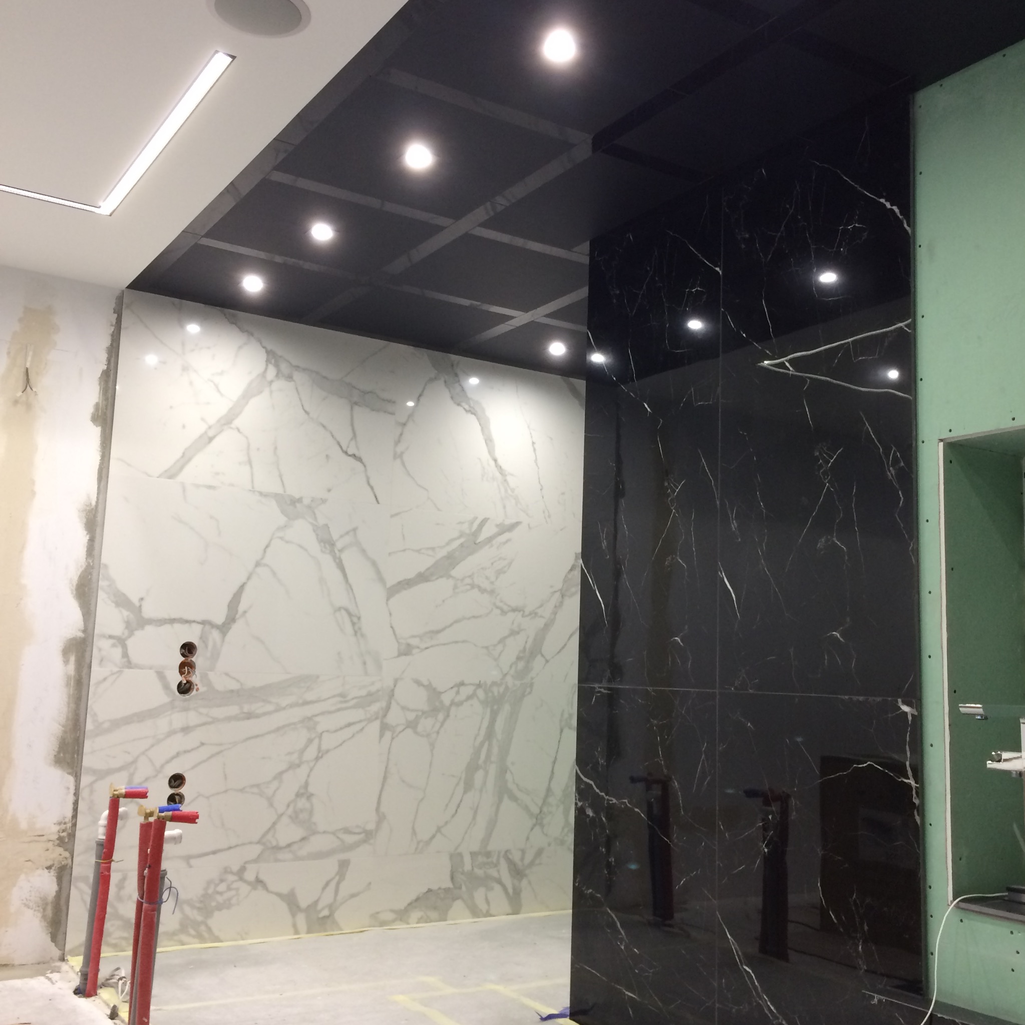 Wnętrze w trakcie prac wykończeniowych: ściany z białych i czarnych płyt imitujących marmur, widoczne instalacje wodne i fragment sufitu z oświetleniem punktowym.