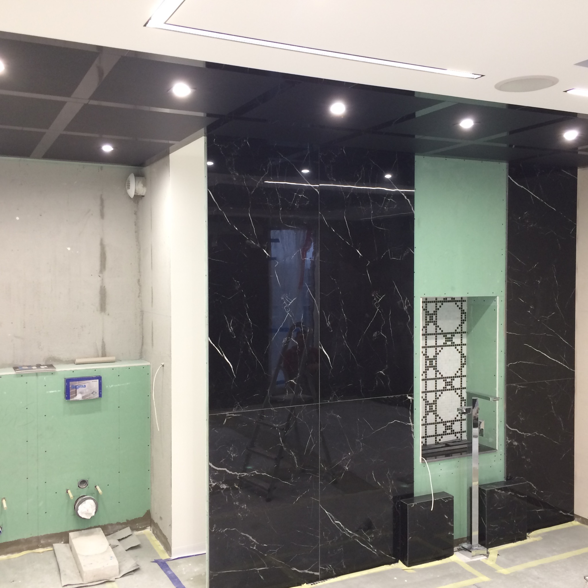 Wnętrze łazienki w trakcie prac wykończeniowych: czarne marmurowe płytki na ścianach, zielona płyta kartonowo-gipsowa z instalacją sanitarną, fragment dekoracyjnej mozaiki w niszy, sufit...