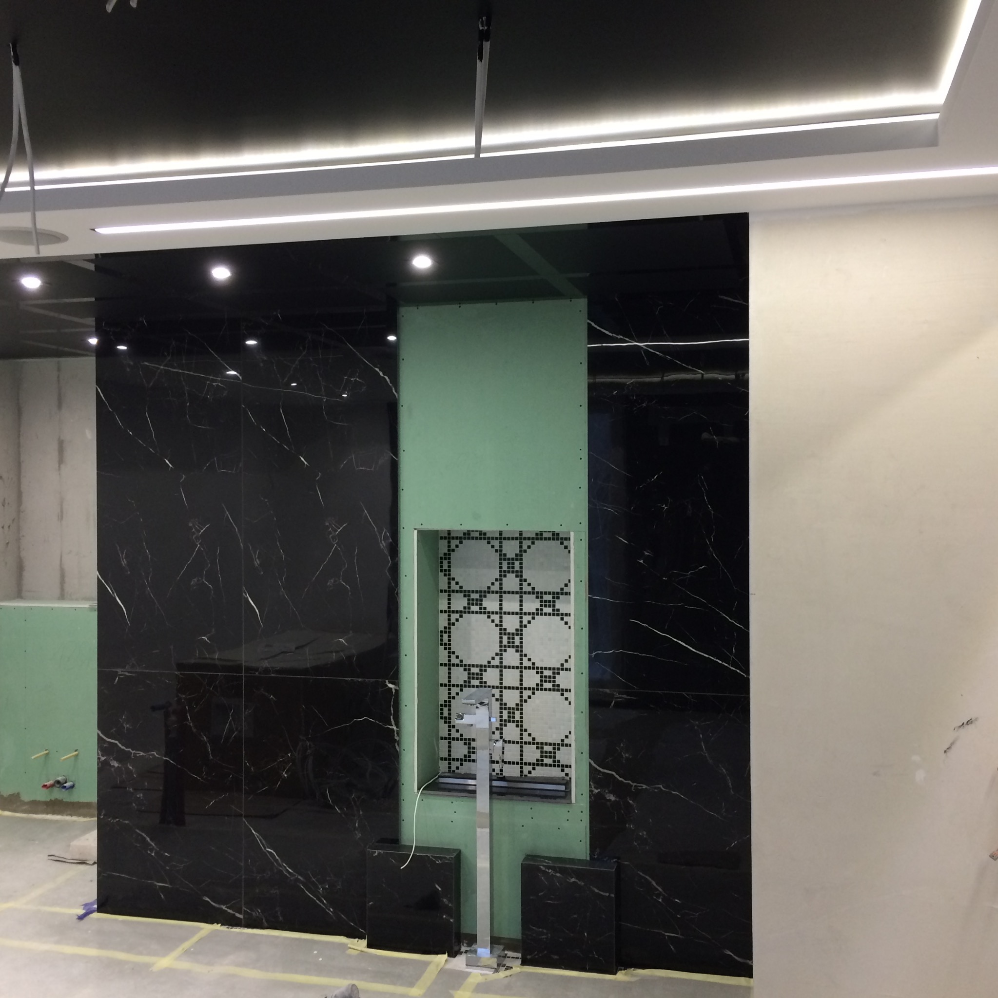 Ściana w trakcie wykańczania z czarnymi płytkami imitującymi marmur, zielona płyta karton-gips z wnęką wyłożoną mozaiką i stojącą baterią umywalkową, widoczne elementy instalacji elektrycznej...