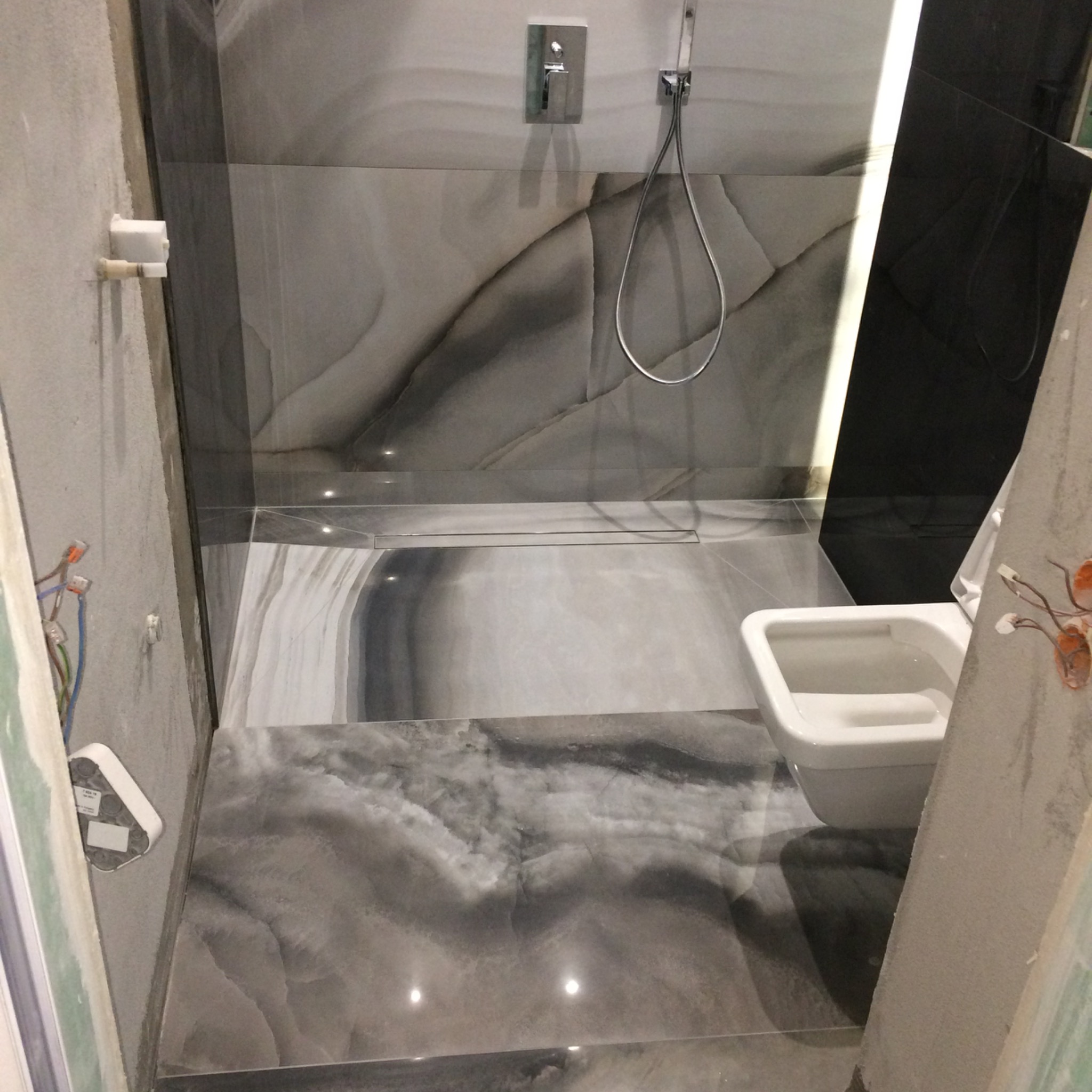 Wnętrze łazienki w trakcie prac wykończeniowych: widoczne szare płytki imitujące marmur na ścianach i podłodze, zamontowana armatura prysznicowa, podtynkowa spłuczka WC oraz surowa ściana...