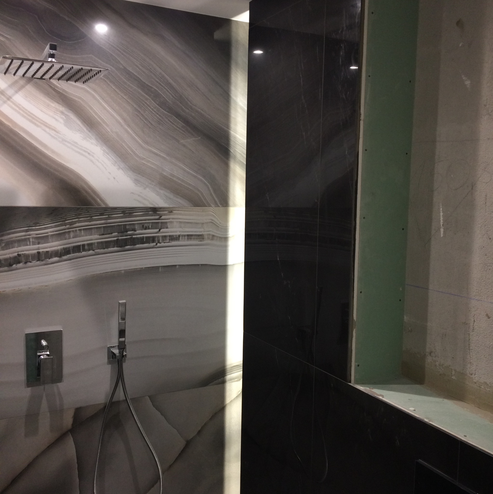 Ściana prysznicowa z panelami imitującymi marmur w odcieniach szarości i bieli, kontrastująca z czarną ścianą obok; widoczna deszczownica i bateria podtynkowa, a także fragment ściany z płytą...
