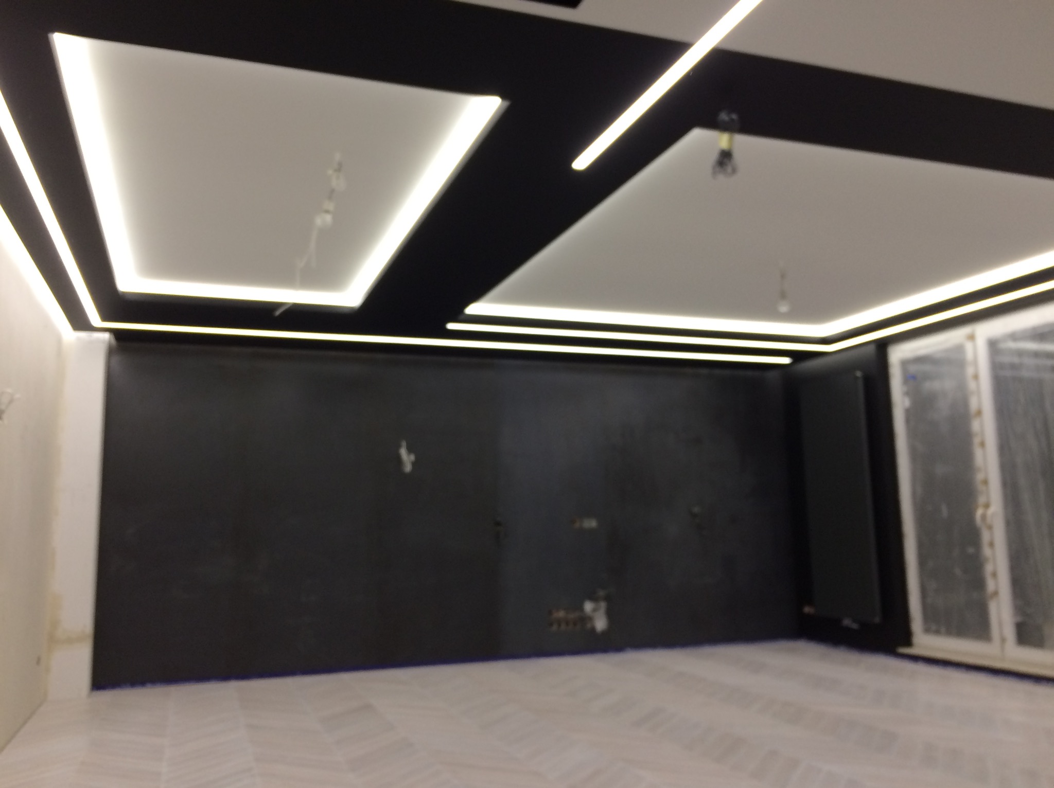 Wnętrze pokoju w trakcie prac wykończeniowych, z czarną ścianą, białymi panelami sufitowymi z oświetleniem LED i podłogą z jasnych płytek imitujących drewno; widoczne niedokończone instalacje...