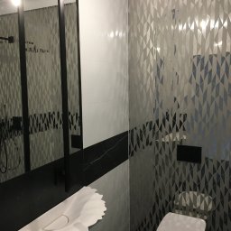 Remonty wykończenia wnętrz Level Art - Nowoczesna łazienka z geometrycznymi płytkami w odcieniach szarości i bieli, unikalna umywalka w kształcie kwiatu, czarne akcenty i minimalistyczna toaleta.