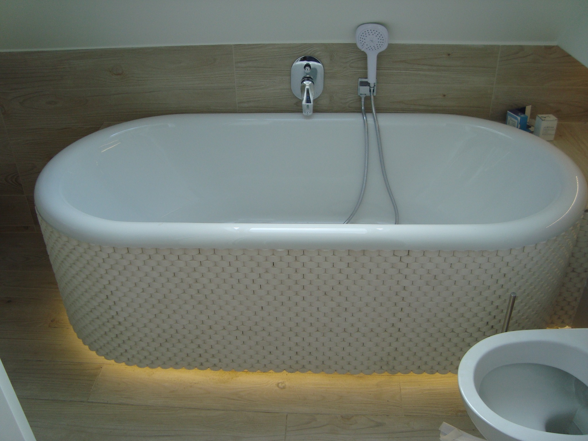 Owalna wanna z dekoracyjną obudową o strukturze plastra miodu, podświetlona od dołu, zamontowana bateria ścienna z słuchawką prysznicową, drewnopodobne płytki na ścianie i podłodze, fragment...