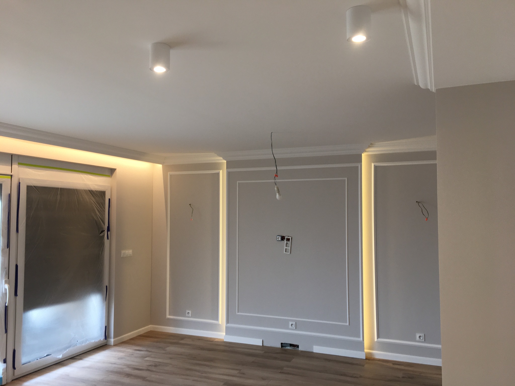 Szare ściany z dekoracyjnymi listwami i oświetleniem LED w nowo wykończonym pokoju, z zabezpieczonymi folią drzwiami balkonowymi i drewnopodobną podłogą, widoczne gniazdka elektryczne i wystające...