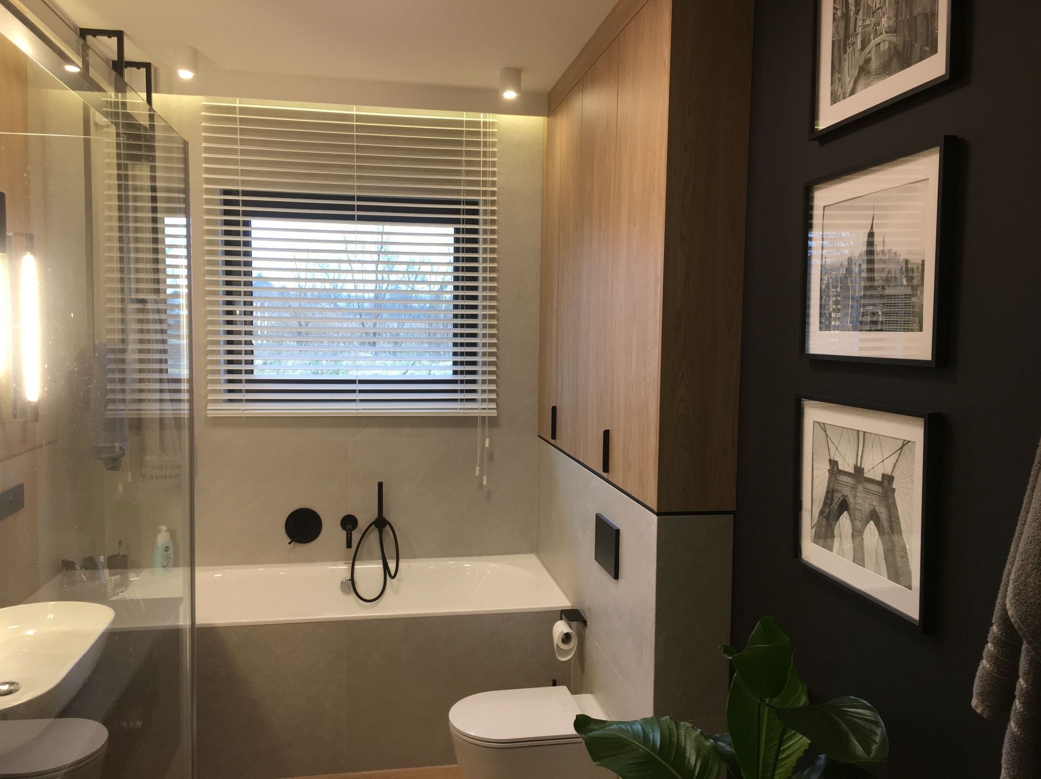 Nowoczesna łazienka z wanną, umywalką nablatową, toaletą, szafkami z drewnianym fornirem, czarną armaturą i trzema czarno-białymi fotografiami na ciemnej ścianie.