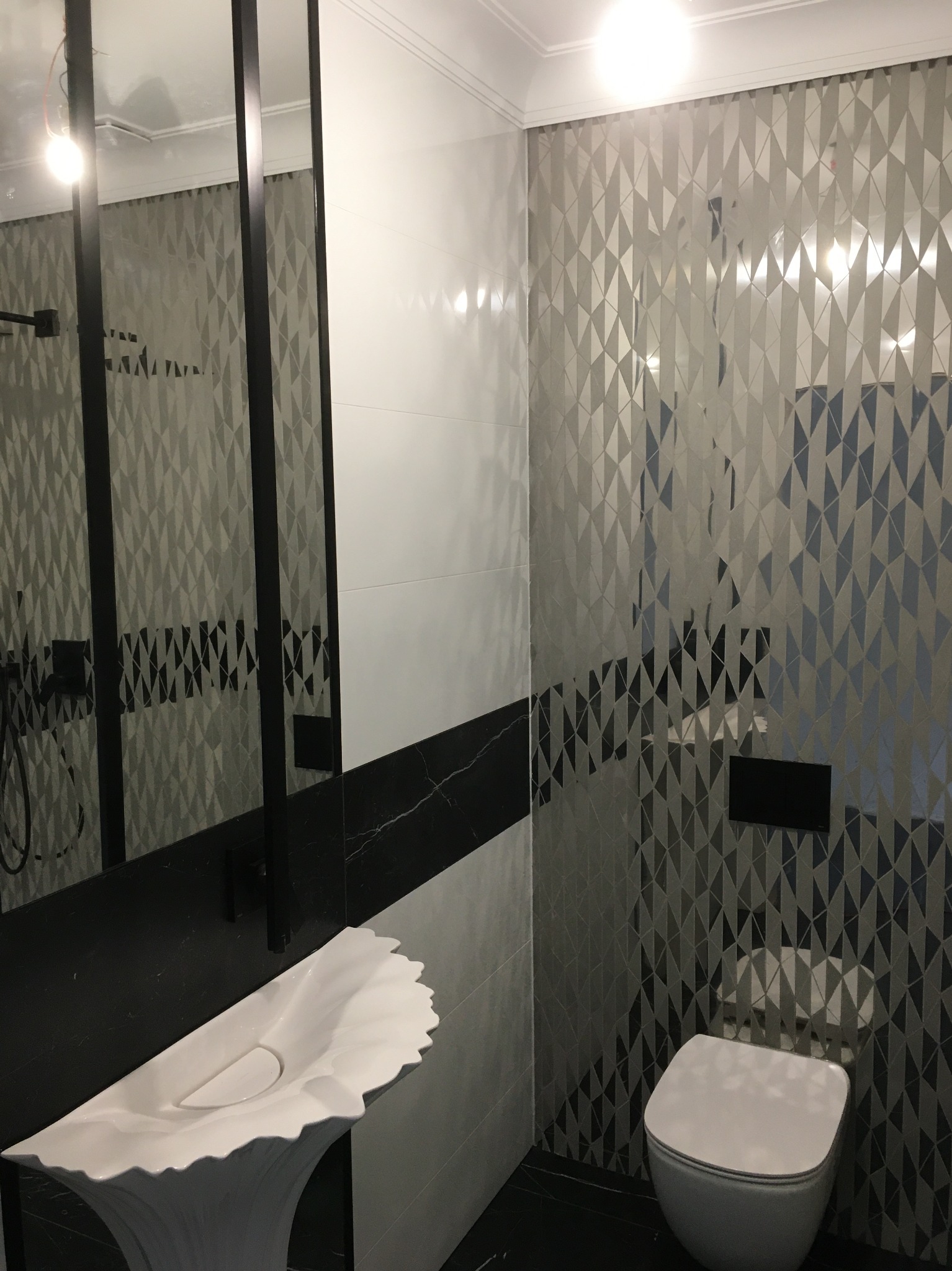 Nowoczesna łazienka z geometrycznymi płytkami w odcieniach szarości i bieli, unikalna umywalka w kształcie kwiatu, czarne akcenty i minimalistyczna toaleta.