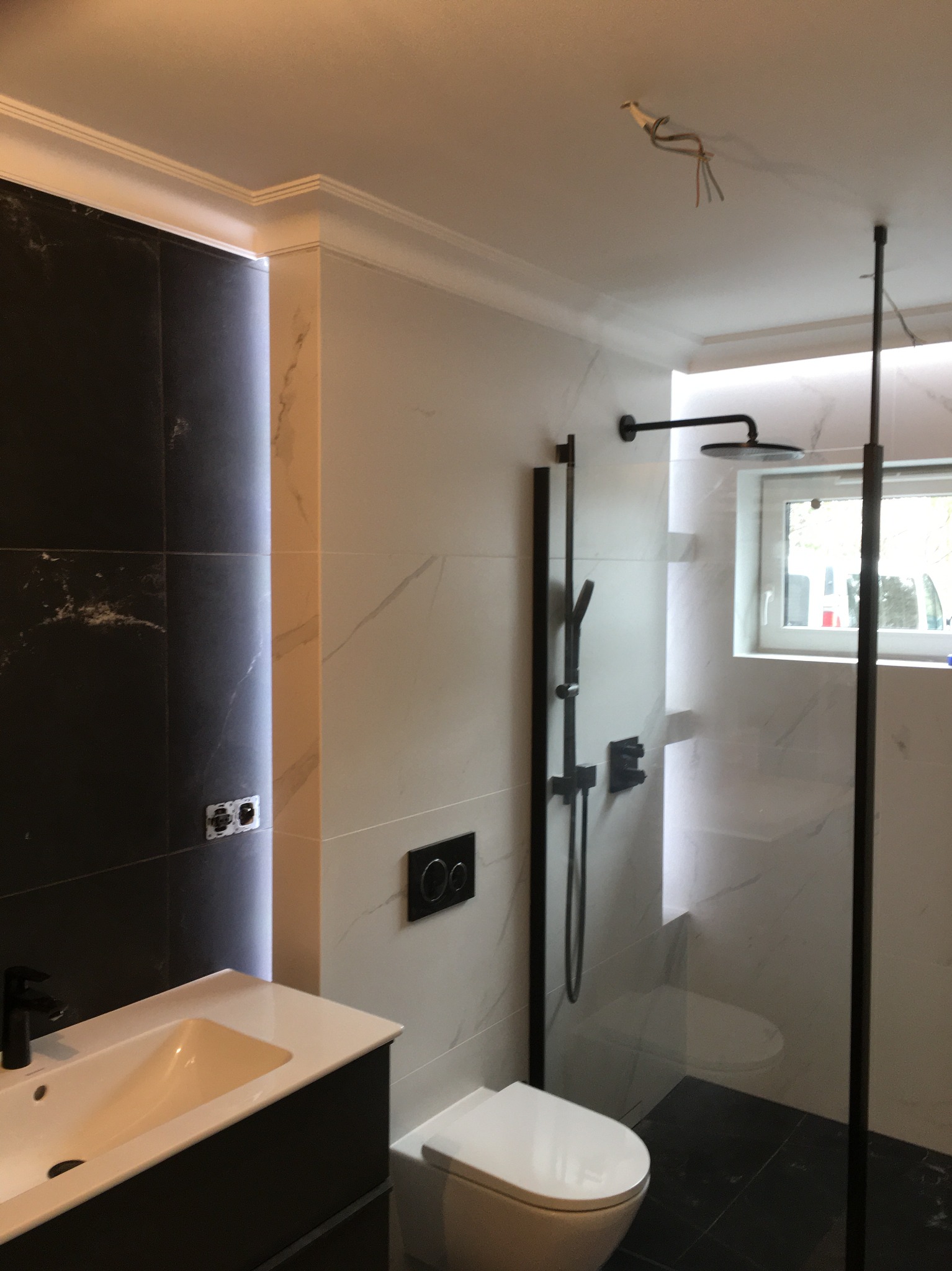 Nowoczesna łazienka z czarno-białymi płytkami marmurkowymi, czarną armaturą, kabiną prysznicową typu walk-in i podwieszaną toaletą, widoczne wystające kable z sufitu.