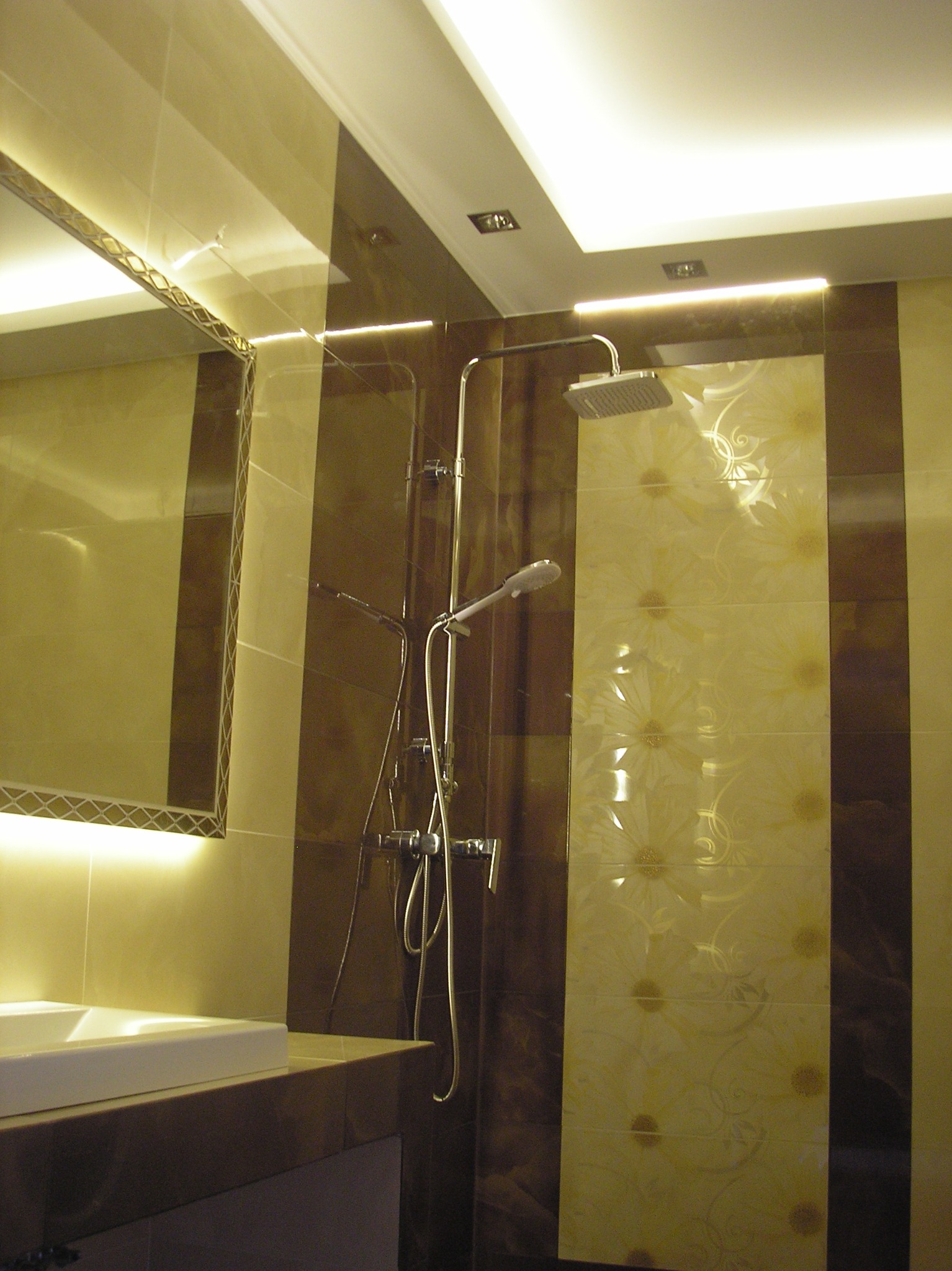 Wykończona łazienka z brązowo-beżowymi płytkami, deszczownicą i podświetlanym lustrem nad umywalką.