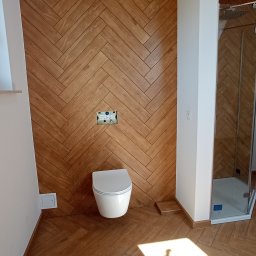 Usługi Ogólnobudowlane - Nowoczesna łazienka z drewnopodobnymi płytkami ułożonymi w jodełkę na ścianie i podłodze, wisząca toaleta oraz kabina prysznicowa z przezroczystego szkła.
