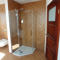 Usługi Ogólnobudowlane - Nowoczesna łazienka z kabiną prysznicową narożną, wykończona drewnopodobną płytką ułożoną w jodełkę na ścianach i podłodze, widoczna również toaleta podwieszana i drewniane drzwi.