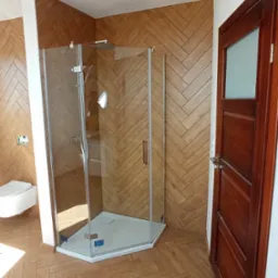 Nowoczesna łazienka z kabiną prysznicową narożną, wykończona drewnopodobną płytką ułożoną w jodełkę na ścianach i podłodze, widoczna również toaleta podwieszana i drewniane drzwi.