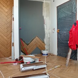 Usługi Ogólnobudowlane - Układanie drewnopodobnej glazury w jodełkę na ścianie i podłodze, widoczne narzędzia glazurnicze, poziomica, puszki po kleju, nieukończone fragmenty ściany, zabezpieczone drzwi folią.