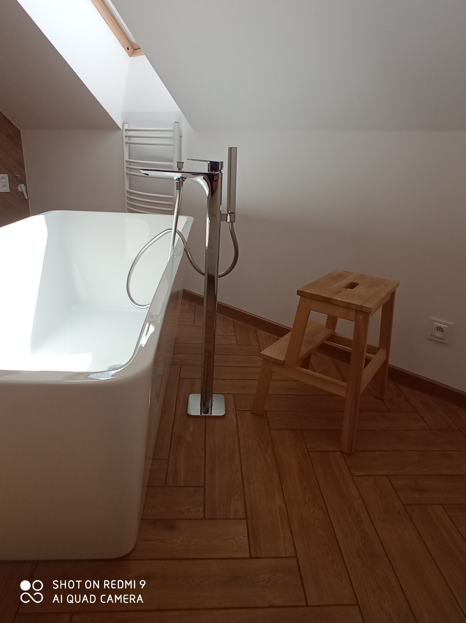 Nowoczesna biała wanna wolnostojąca z chromowaną armaturą na tle poddasza z drewnianą podłogą i drabinką grzejnikową.