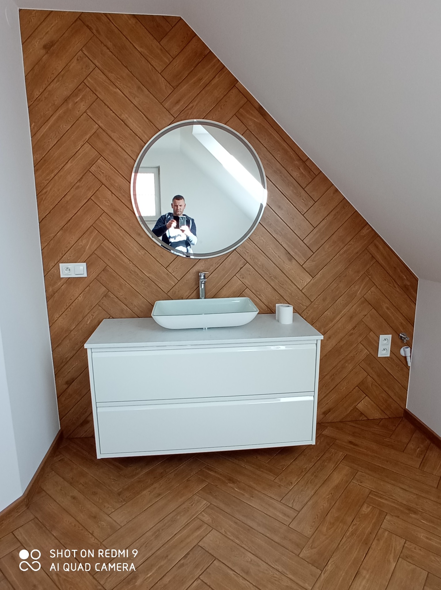 Łazienka na poddaszu z drewnopodobną okładziną w jodełkę na ścianach i podłodze, biała szafka z umywalką nablatową i okrągłym lustrem, w którym odbija się osoba robiąca zdjęcie.