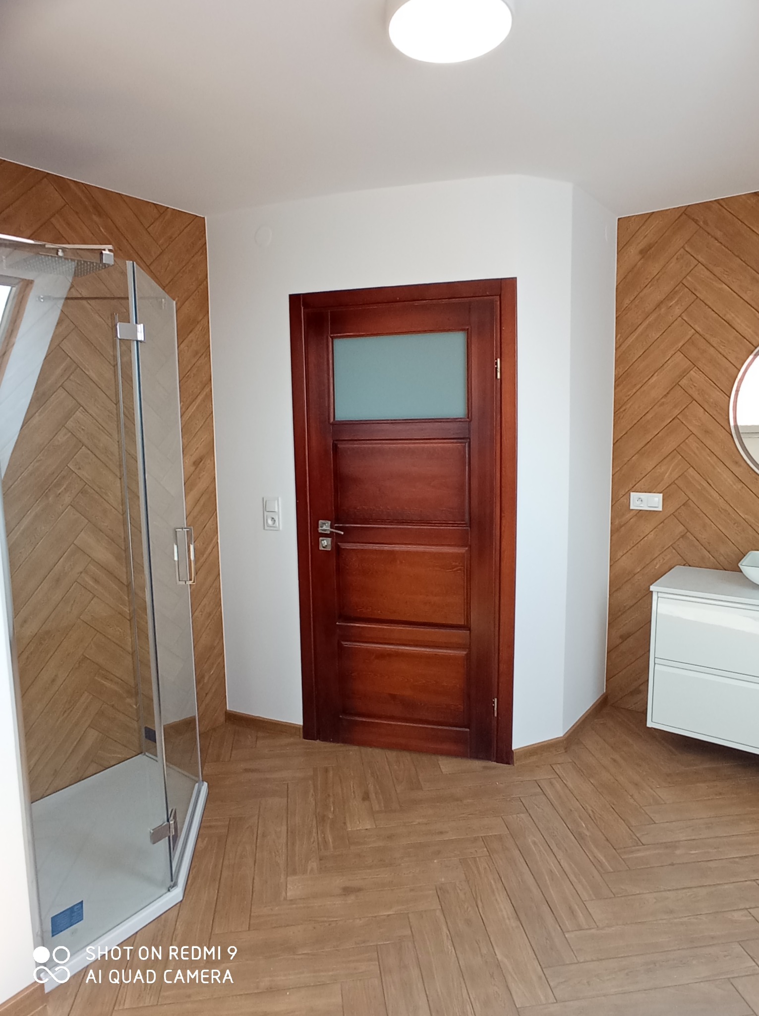 Nowoczesna łazienka z kabiną prysznicową, drewnianą podłogą i ścianą w jodełkę, ciemne drewniane drzwi z szybą i biała szafka z umywalką.