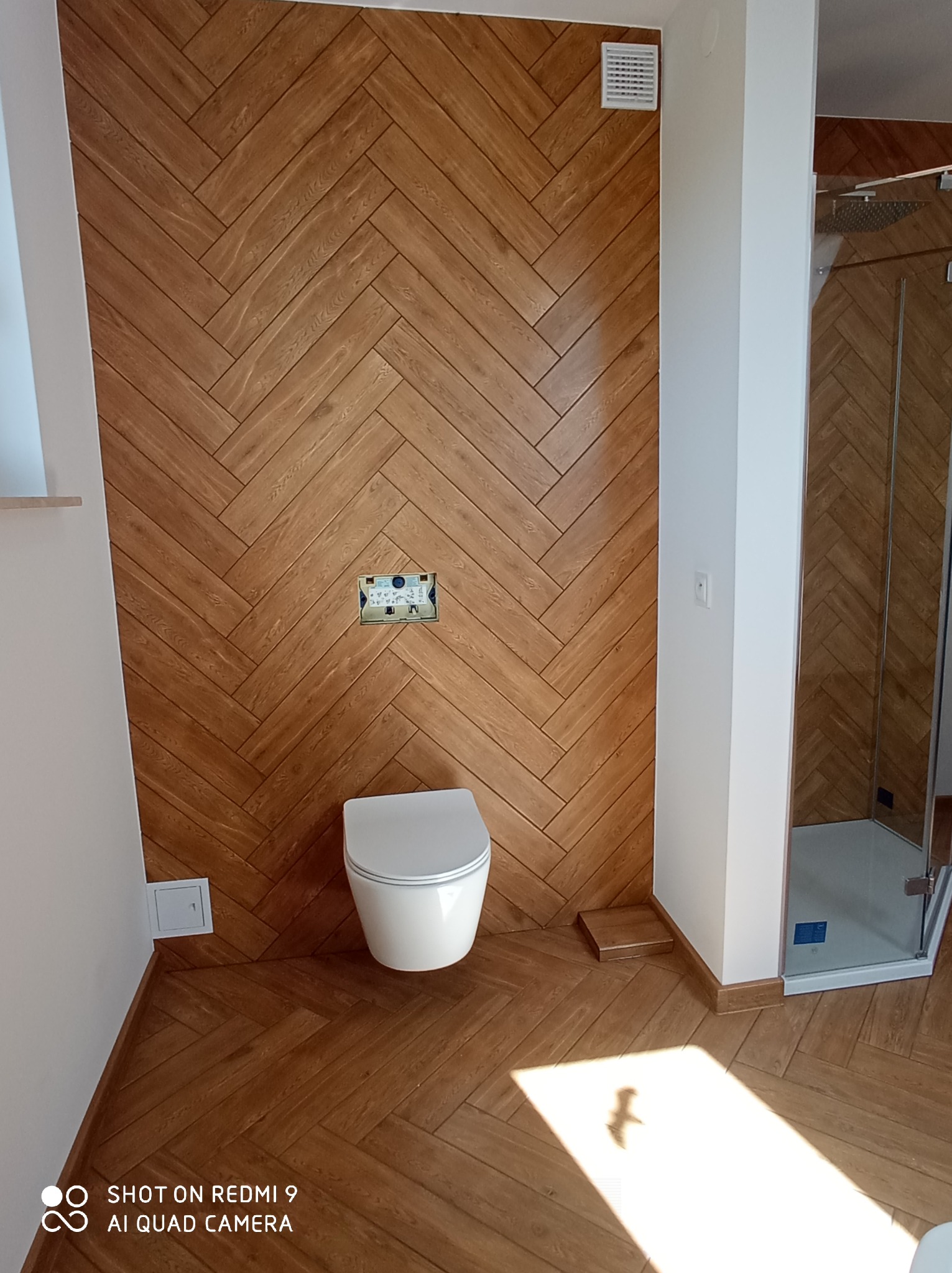 Nowoczesna łazienka z drewnopodobnymi płytkami ułożonymi w jodełkę na ścianie i podłodze, wisząca toaleta oraz kabina prysznicowa z przezroczystego szkła.