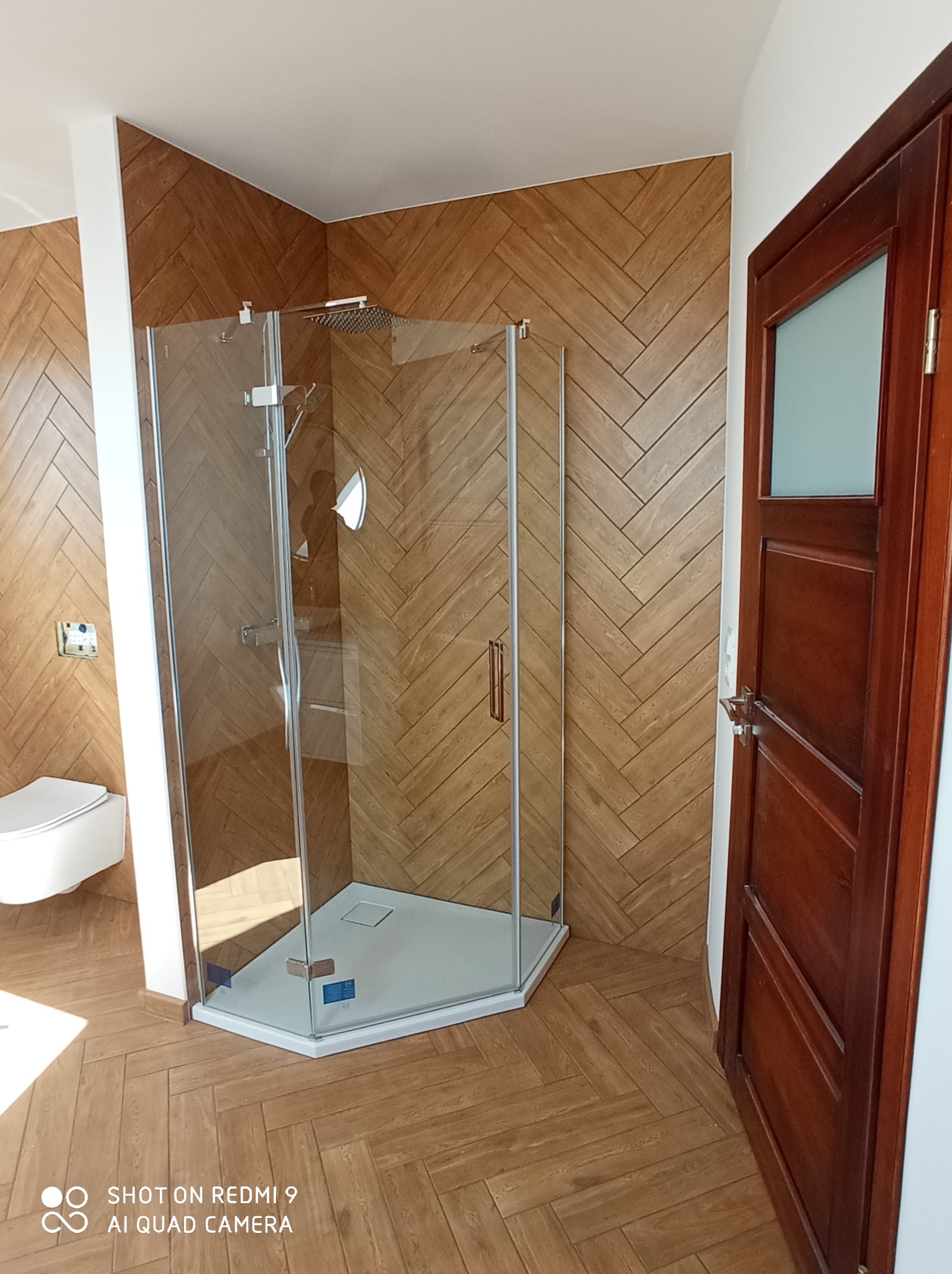 Nowoczesna łazienka z kabiną prysznicową narożną, wykończona drewnopodobną płytką ułożoną w jodełkę na ścianach i podłodze, widoczna również toaleta podwieszana i drewniane drzwi.