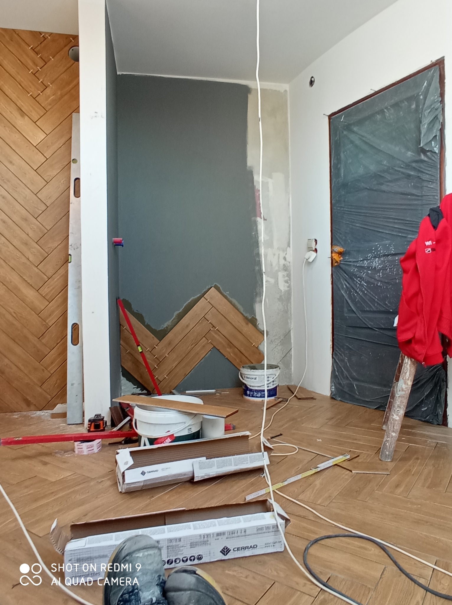 Układanie drewnopodobnej glazury w jodełkę na ścianie i podłodze, widoczne narzędzia glazurnicze, poziomica, puszki po kleju, nieukończone fragmenty ściany, zabezpieczone drzwi folią.