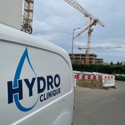 Hydro-Clinique Mateusz Grochalak - Biały van z logo 'Hydro Clinique' na tle budowy z żurawiami. Logo w kształcie kropli wody. Widok z boku, częściowo na chodniku z kostki.