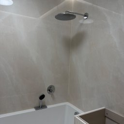 Hydro-Clinique Mateusz Grochalak - Nowoczesna łazienka z wanną, deszczownicą i beżowymi płytkami. Widoczny fragment szafki pod umywalkę z ryflowanym frontem. Oświetlenie LED w górnej części.