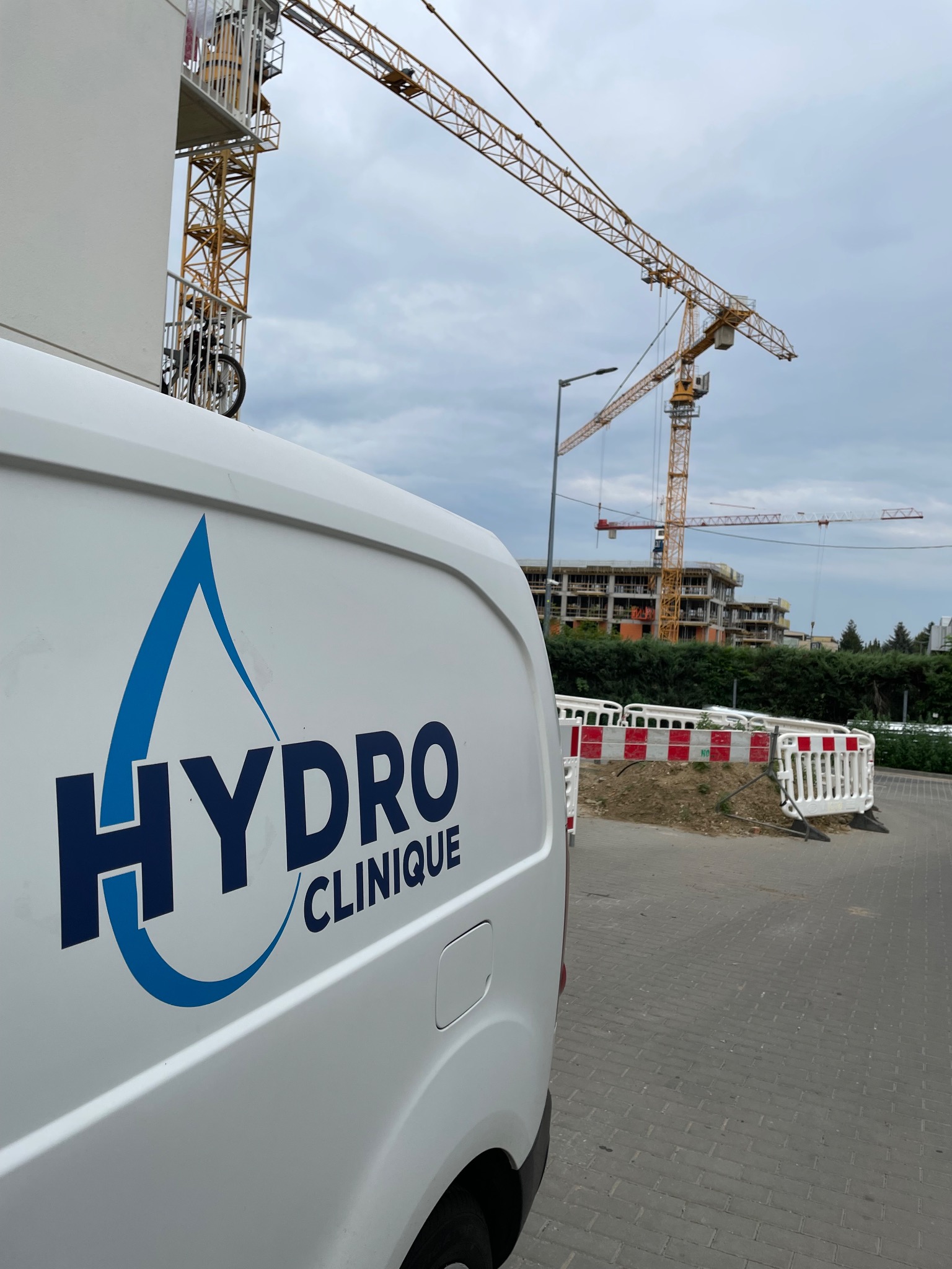 Biały van z logo 'Hydro Clinique' na tle budowy z żurawiami. Logo w kształcie kropli wody. Widok z boku, częściowo na chodniku z kostki.
