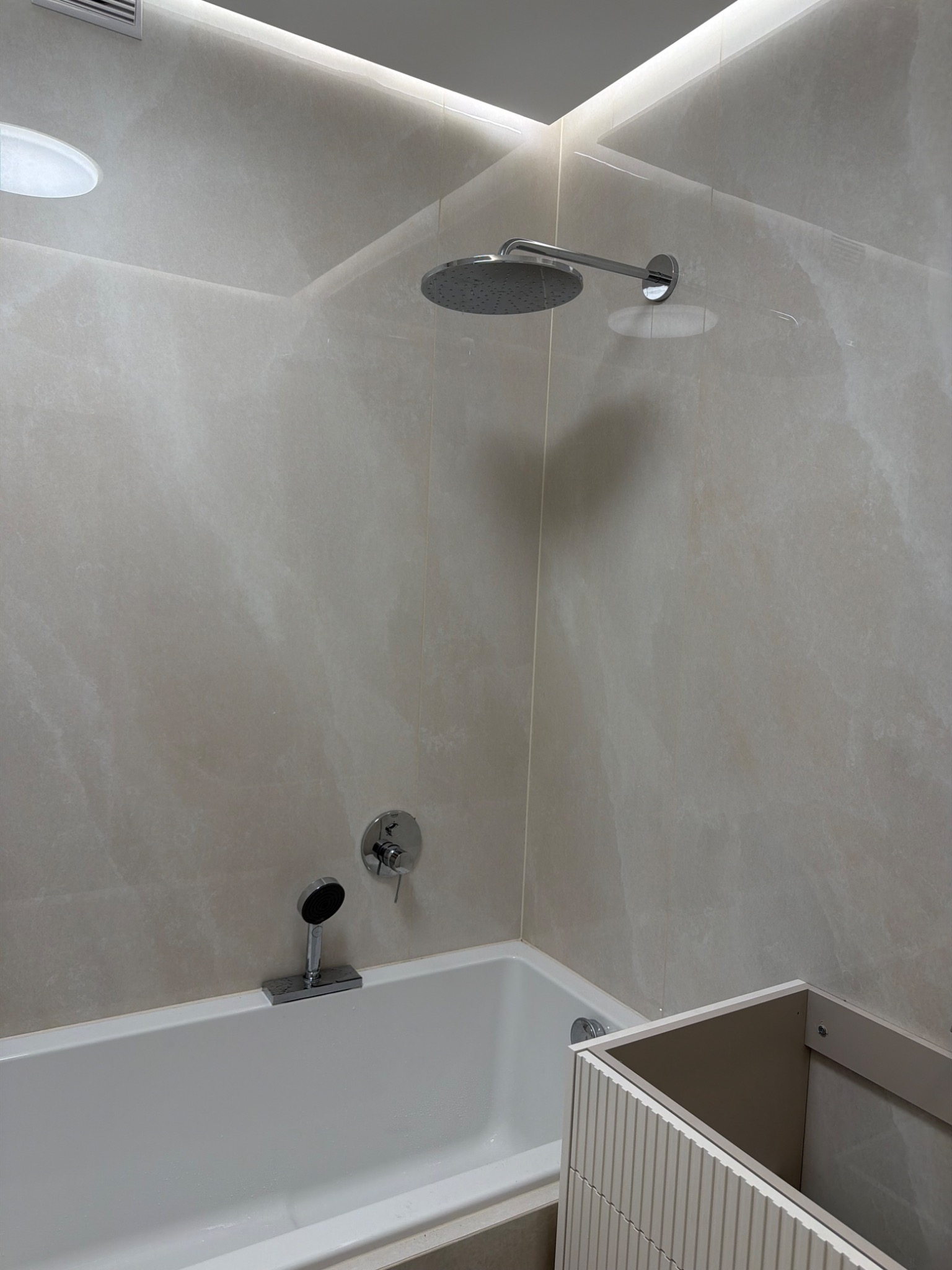 Nowoczesna łazienka z wanną, deszczownicą i beżowymi płytkami. Widoczny fragment szafki pod umywalkę z ryflowanym frontem. Oświetlenie LED w górnej części.