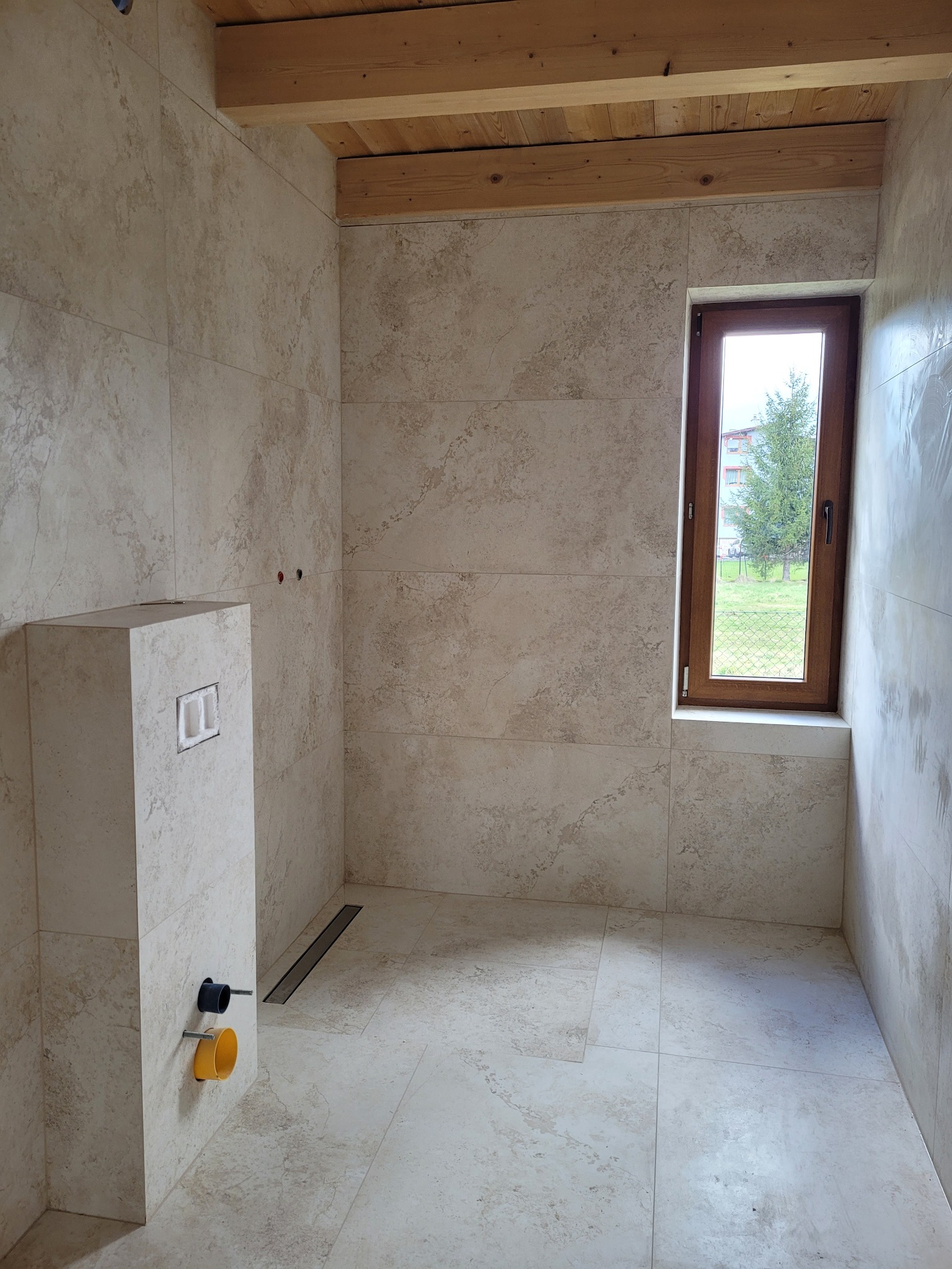 Jasna, wykafelkowana łazienka z drewnianym stropem, oknem z widokiem na zieleń, zabudową WC i odpływem liniowym w podłodze.