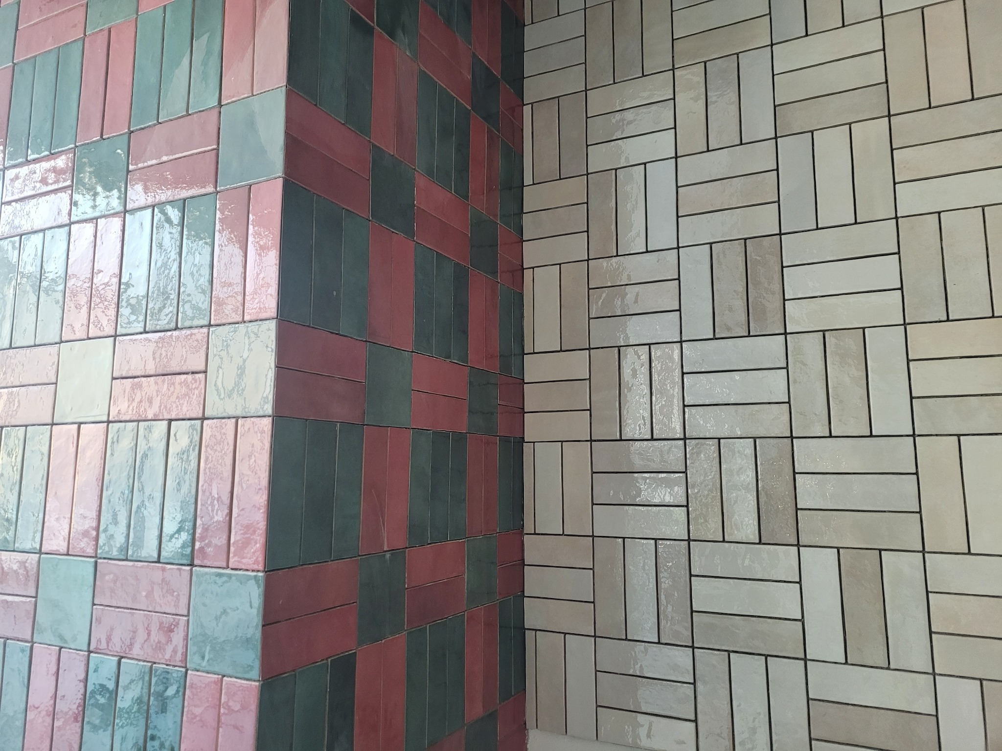 Ściana wyłożona błyszczącymi, prostokątnymi płytkami w dwóch różnych wzorach: po lewej stronie mozaika z płytek w odcieniach różu, zieleni i szarości, po prawej stronie wzór z beżowych płytek...