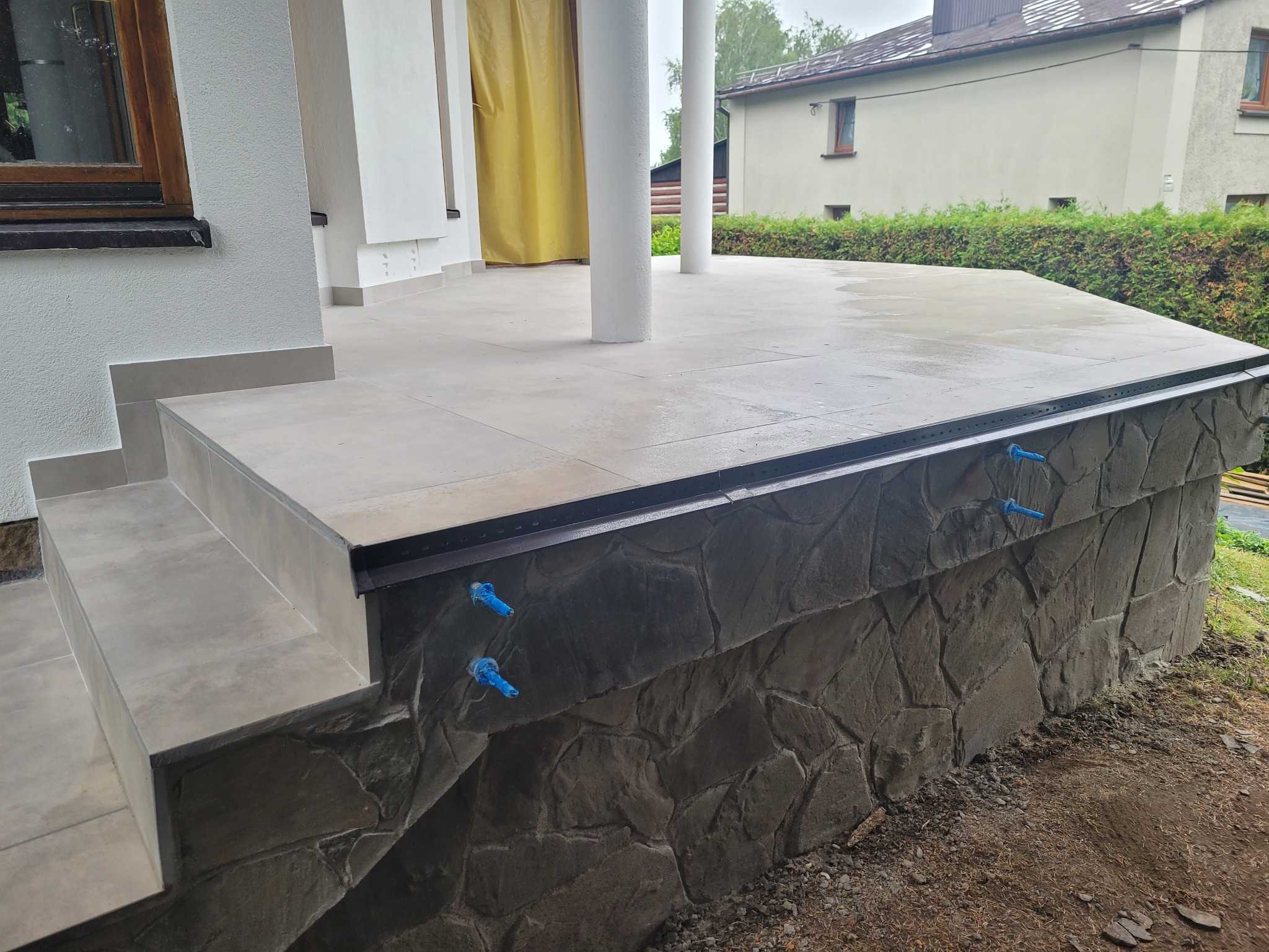 Wykończony taras z kamiennymi schodami w Grodkowie, z widocznymi niebieskimi zaślepkami na ścianie z kamienia dekoracyjnego.