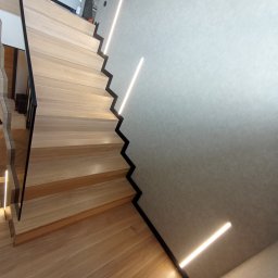 STYL DREAM HOME SPÓŁKA Z OGRANICZONĄ ODPOWIEDZIALNOŚCIĄ - Drewniane schody z czarnymi detalami i oświetleniem LED wzdłuż ściany, widoczne narzędzia i tuba uszczelniająca na podłodze.