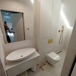 STYL DREAM HOME SPÓŁKA Z OGRANICZONĄ ODPOWIEDZIALNOŚCIĄ - Nowoczesna łazienka w jasnych odcieniach z ryflowanymi szafkami, owalną umywalką nablatową, złotą armaturą i widocznym odbiciem osoby robiącej zdjęcie w lustrze.