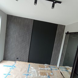 STYL DREAM HOME SPÓŁKA Z OGRANICZONĄ ODPOWIEDZIALNOŚCIĄ - Montaż paneli lamelowych na ścianie w trakcie prac wykończeniowych, zabezpieczona podłoga, czarna ściana w tle, widoczne oświetlenie sufitowe.