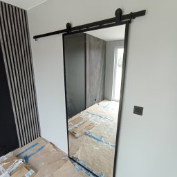 STYL DREAM HOME SPÓŁKA Z OGRANICZONĄ ODPOWIEDZIALNOŚCIĄ - Montaż przesuwnych drzwi z czarną ramą i szyną w pomieszczeniu z panelami ściennymi, widoczne zabezpieczenia podłogi i elementy wykończeniowe.
