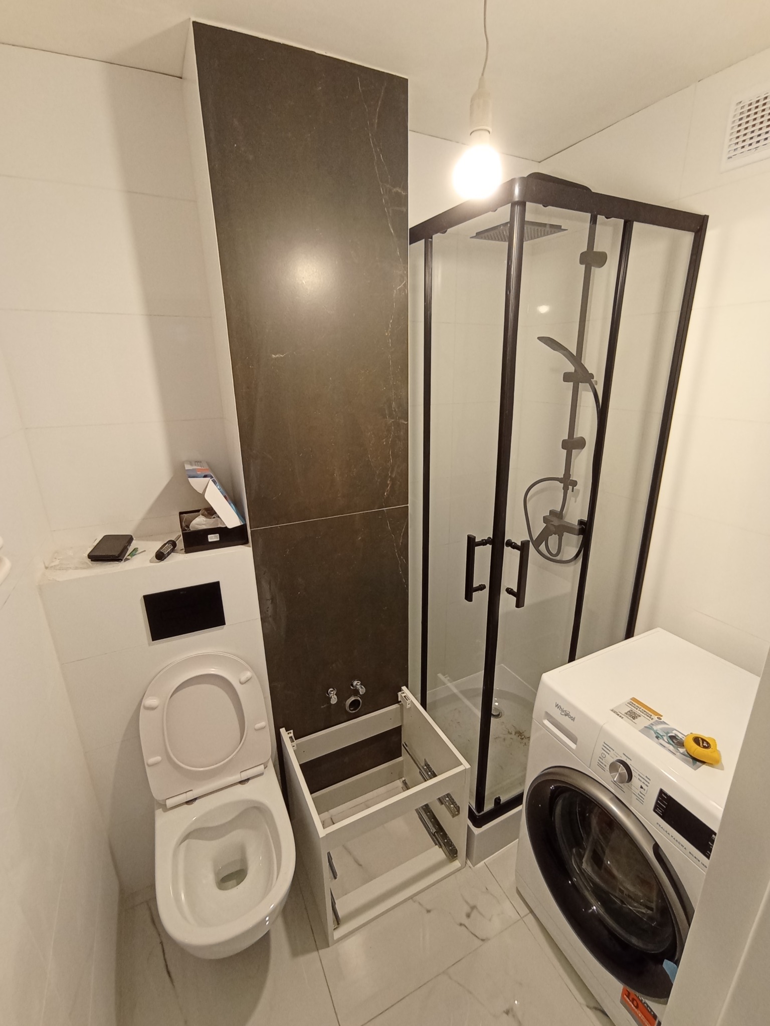 Niedokończona aranżacja łazienki z kabiną prysznicową z czarną ramą, pralką Whirlpool i podwieszaną toaletą z otwartą deską, obok szafka pod umywalkę bez frontów.