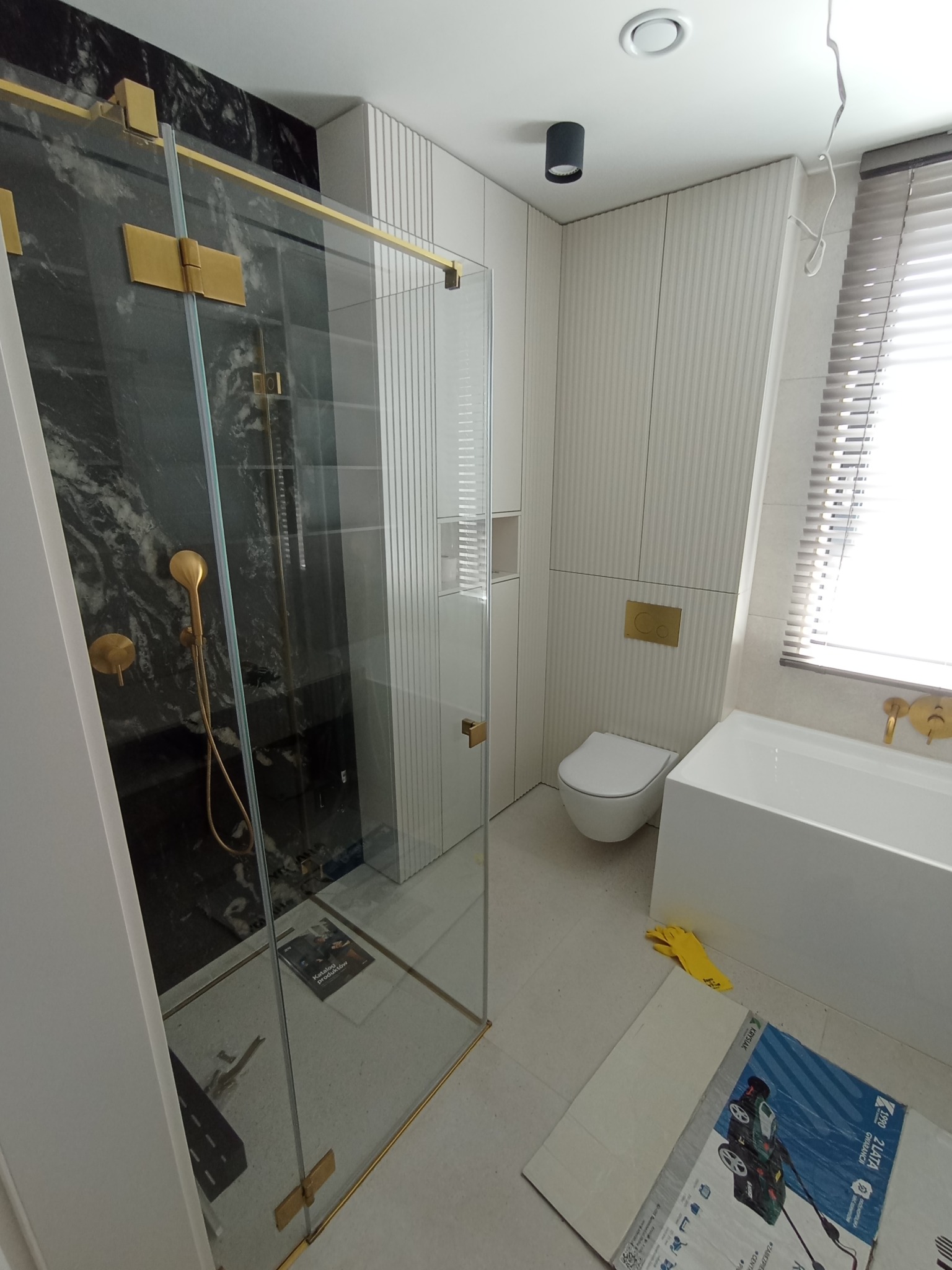 Nowoczesna łazienka z kabiną prysznicową ze złotymi elementami, czarnym marmurem na ścianie, białą armaturą i żółtymi rękawicami leżącymi na podłodze obok opakowania produktu.