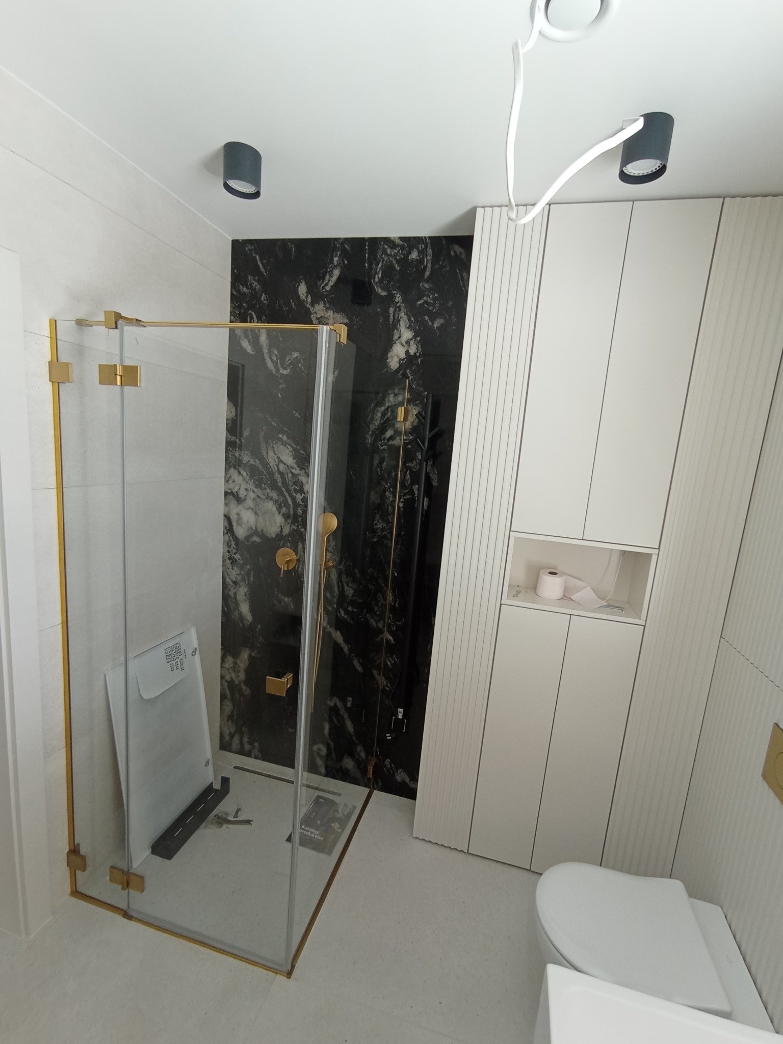 Nowoczesna łazienka z kabiną prysznicową ze złotymi okuciami na tle ściany z czarnego marmuru, biała szafka z toaletą i designerskie oświetlenie sufitowe.