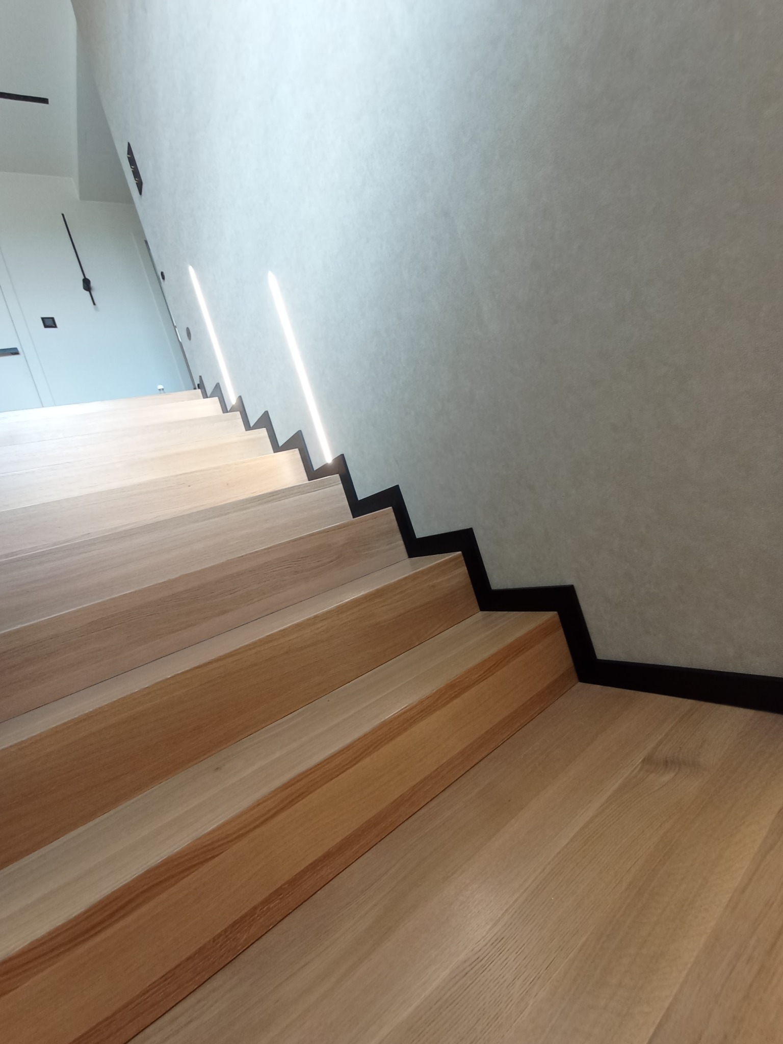 Nowoczesne drewniane schody z czarnym wykończeniem przy ścianie oświetlone subtelnymi pionowymi lampami.