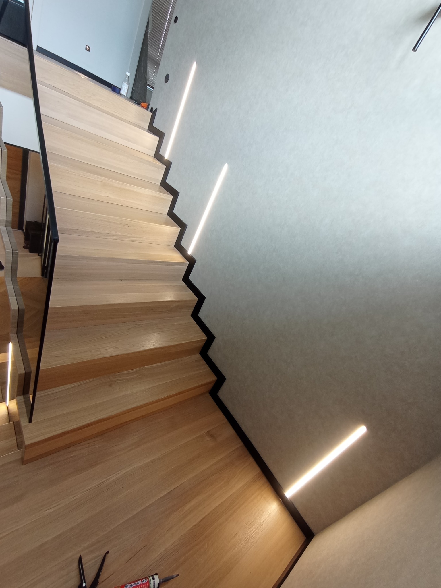 Drewniane schody z czarnymi detalami i oświetleniem LED wzdłuż ściany, widoczne narzędzia i tuba uszczelniająca na podłodze.